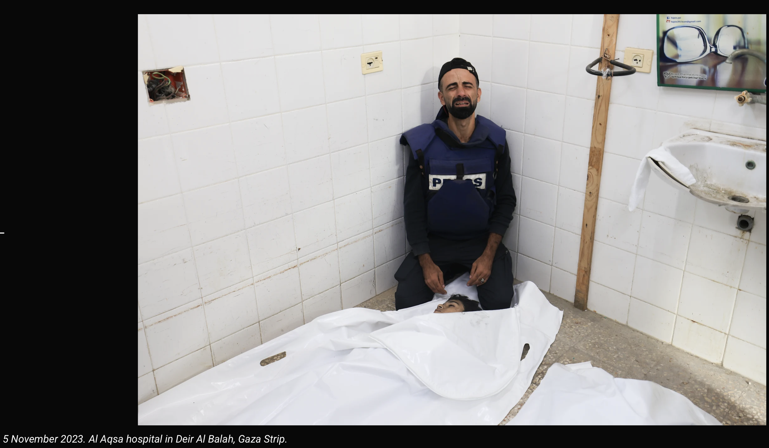 A man in a police vest and cap kneels beside a person lying on the ground, wrapped in a white sheet in a tiled room with a sink and electrical outlets. The room appears to be a hospital or morgue. The photo is dated November 5, 2023, at Al Aqsa Hospi