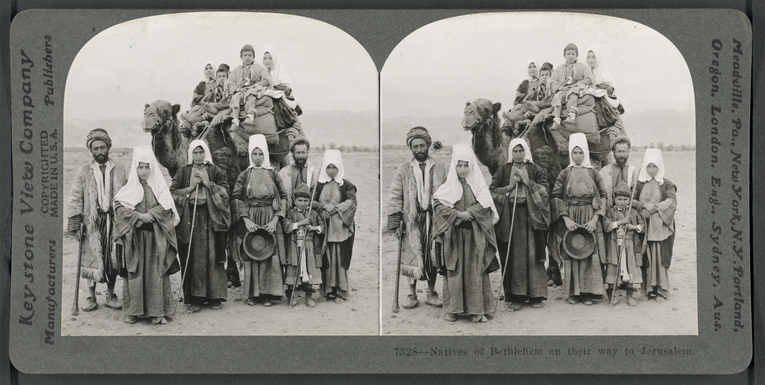 Black-and-white photograph of a group of people, including men, women, and children, standing outside with a camel carrying people in the background. The camel is loaded with passengers and supplies. The setting appears to be a plain or desert landsc