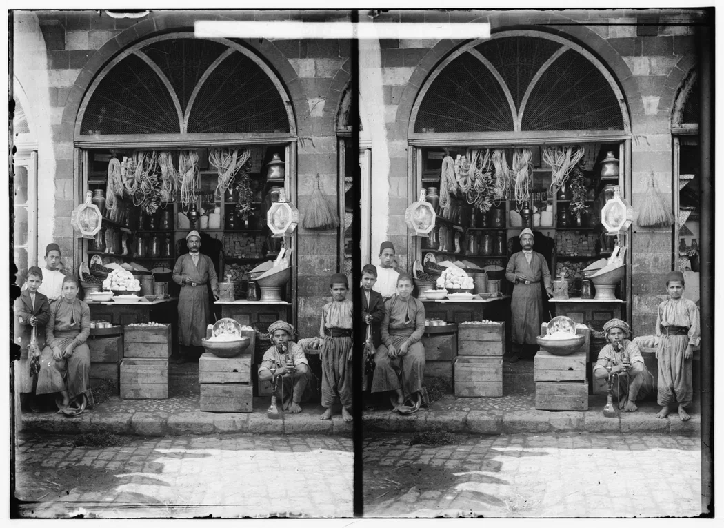 Vintage black and white photograph of a street vendor stand with children and an adult, showing tools and goods for sale in an arched storefront.