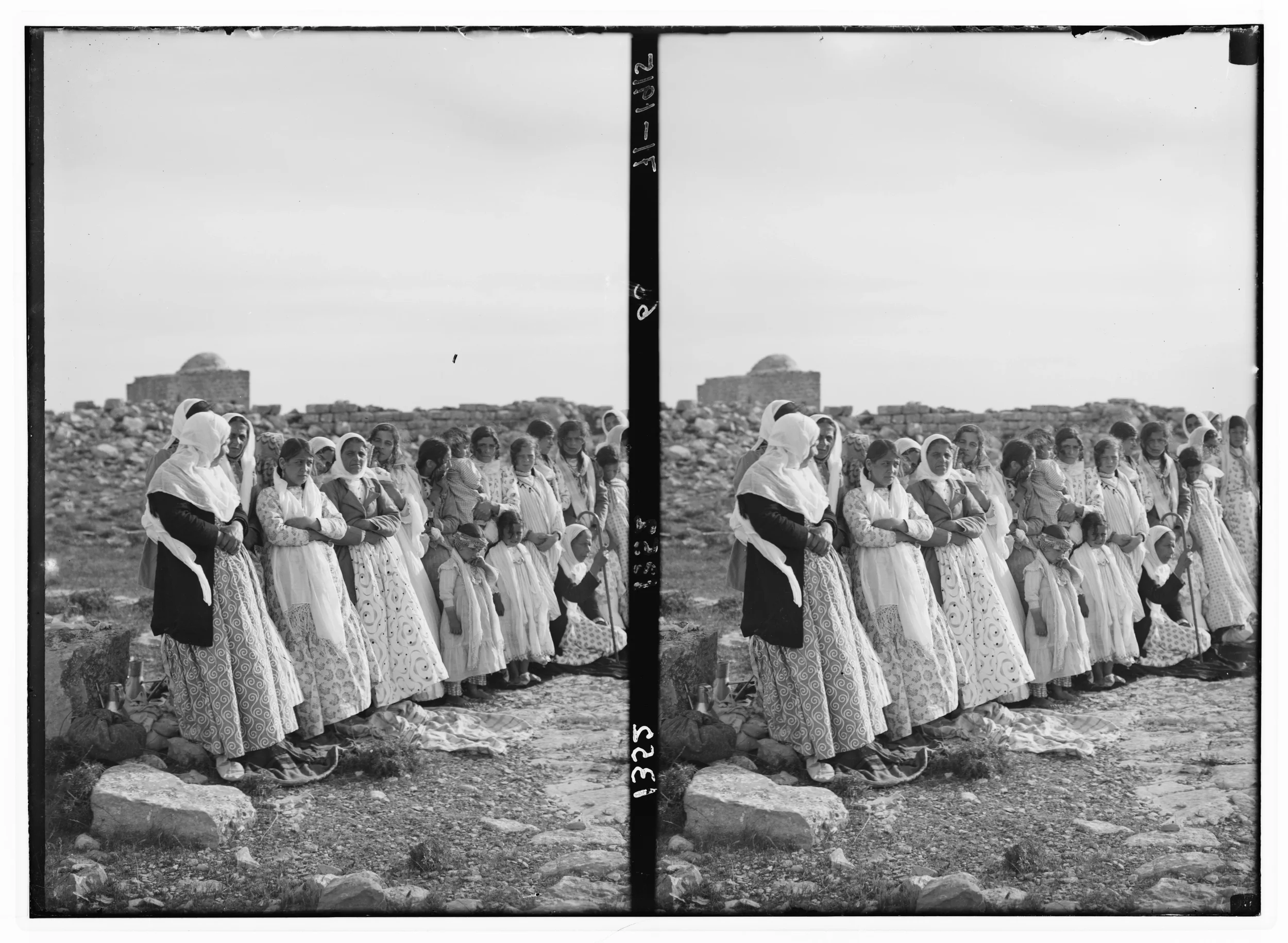 Black and white photograph of a group of women and children standing outdoors on rocky ground, wearing traditional clothing, with a building or structure and open sky in the background.