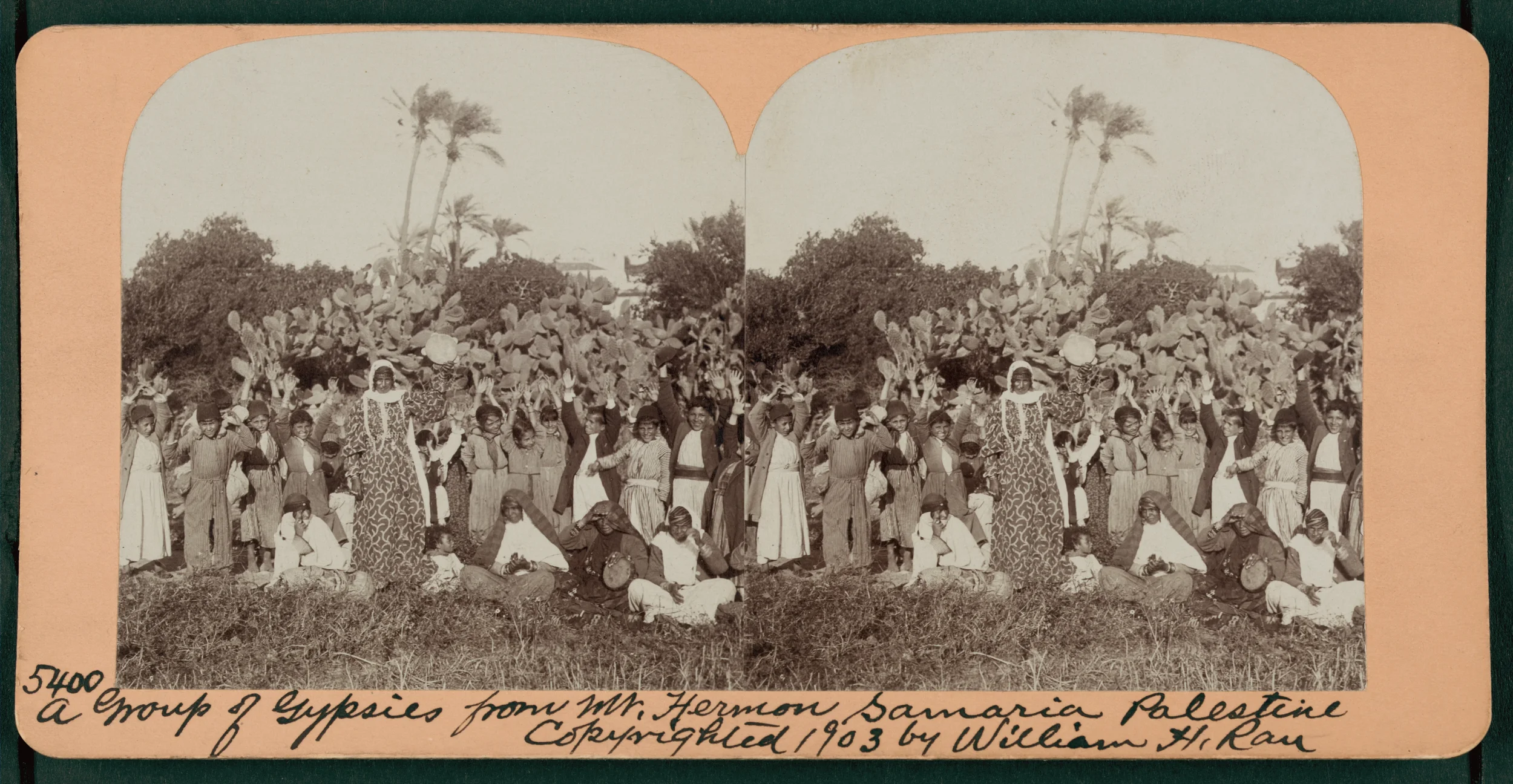 A vintage black-and-white photo showing a group of children and adults outdoors among cactus plants and trees, with some palm trees in the background. The people are smiling and waving at the camera. The photo appears to be historical, with individua