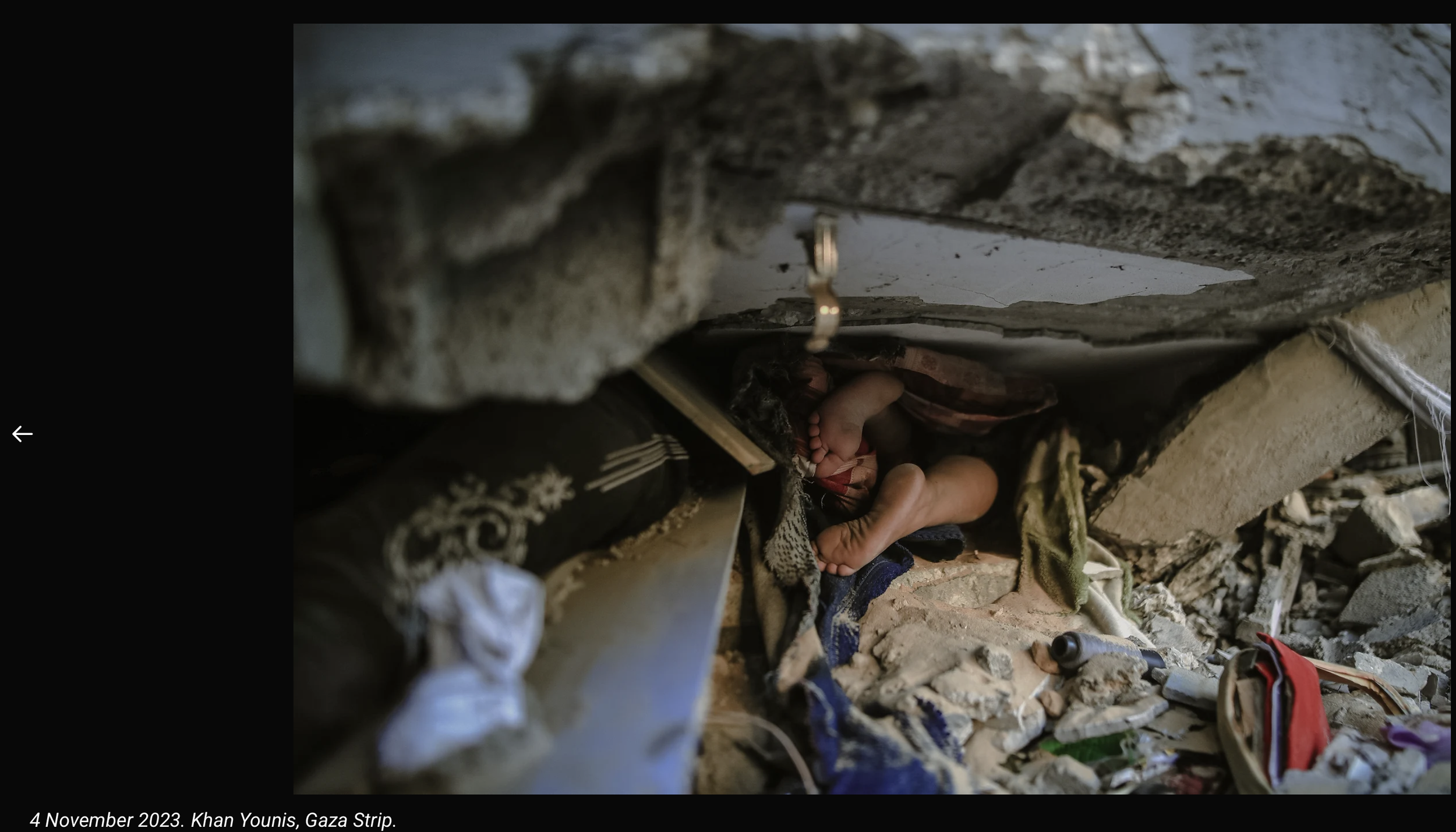 A person lying underneath a rubble-filled area, possibly repairing or inspecting the underneath of a structure. The surrounding area is cluttered with debris and objects.