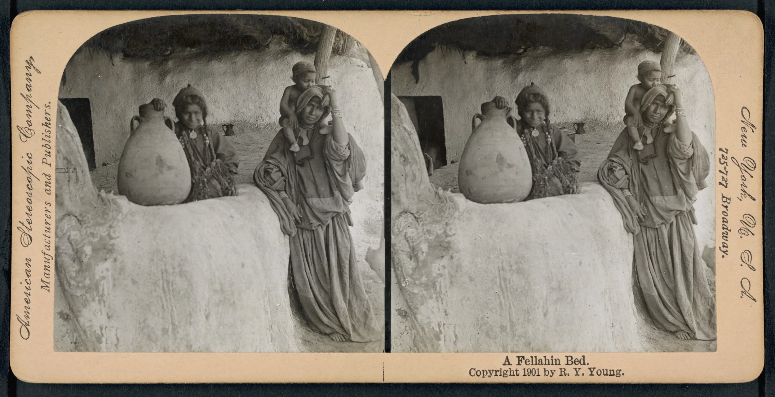A black and white stereoscopic photograph of two women and a child, sitting outside a mud hut. The women are dressed in traditional clothing with jewelry, and the child is sitting on one woman's shoulder. There is a large clay pot on the ledge in fro