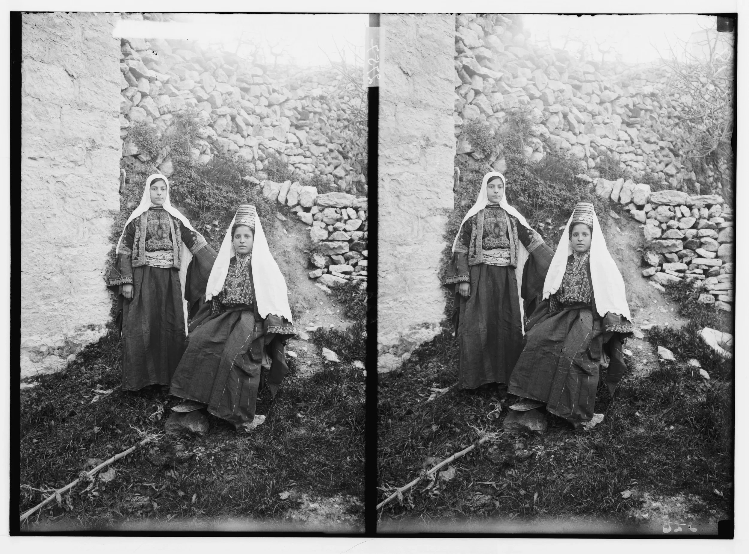 A black and white photograph of two young girls in traditional clothing, standing outdoors against a stone wall and rocky terrain. Both girls are looking directly at the camera with serious expressions.