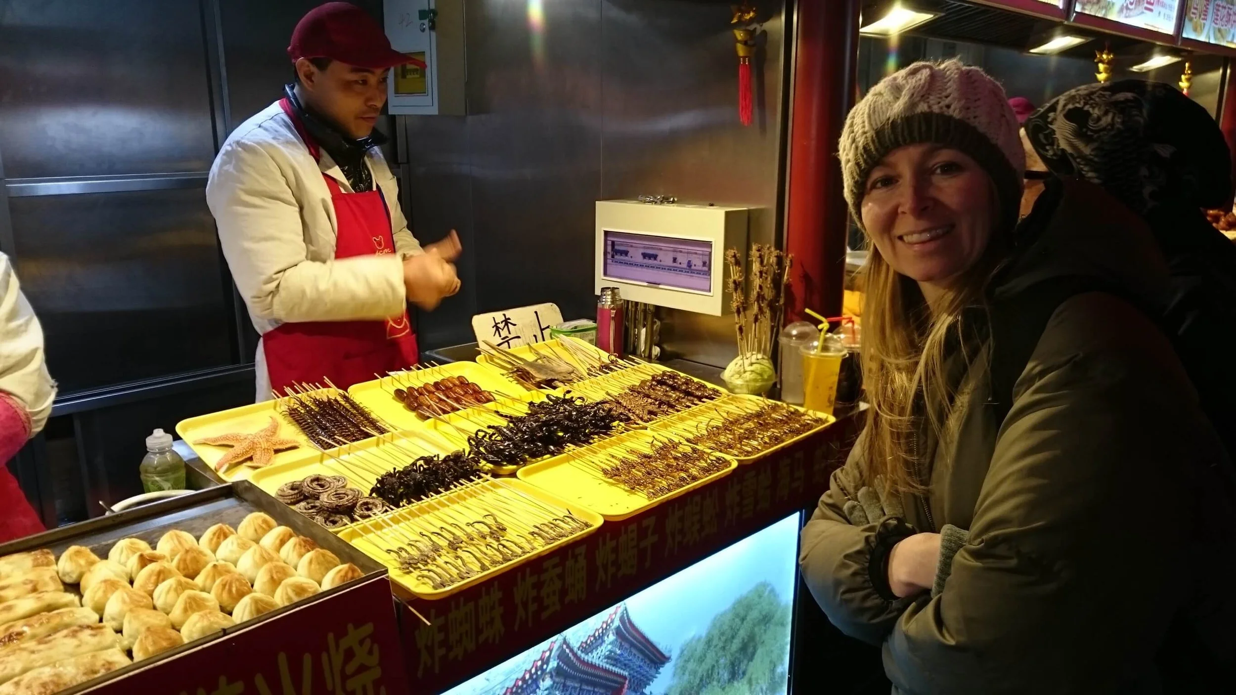 A woman smiling at a street food stall selling skewered insects and other snacks, with a vendor behind the counter and a display of food items in front.