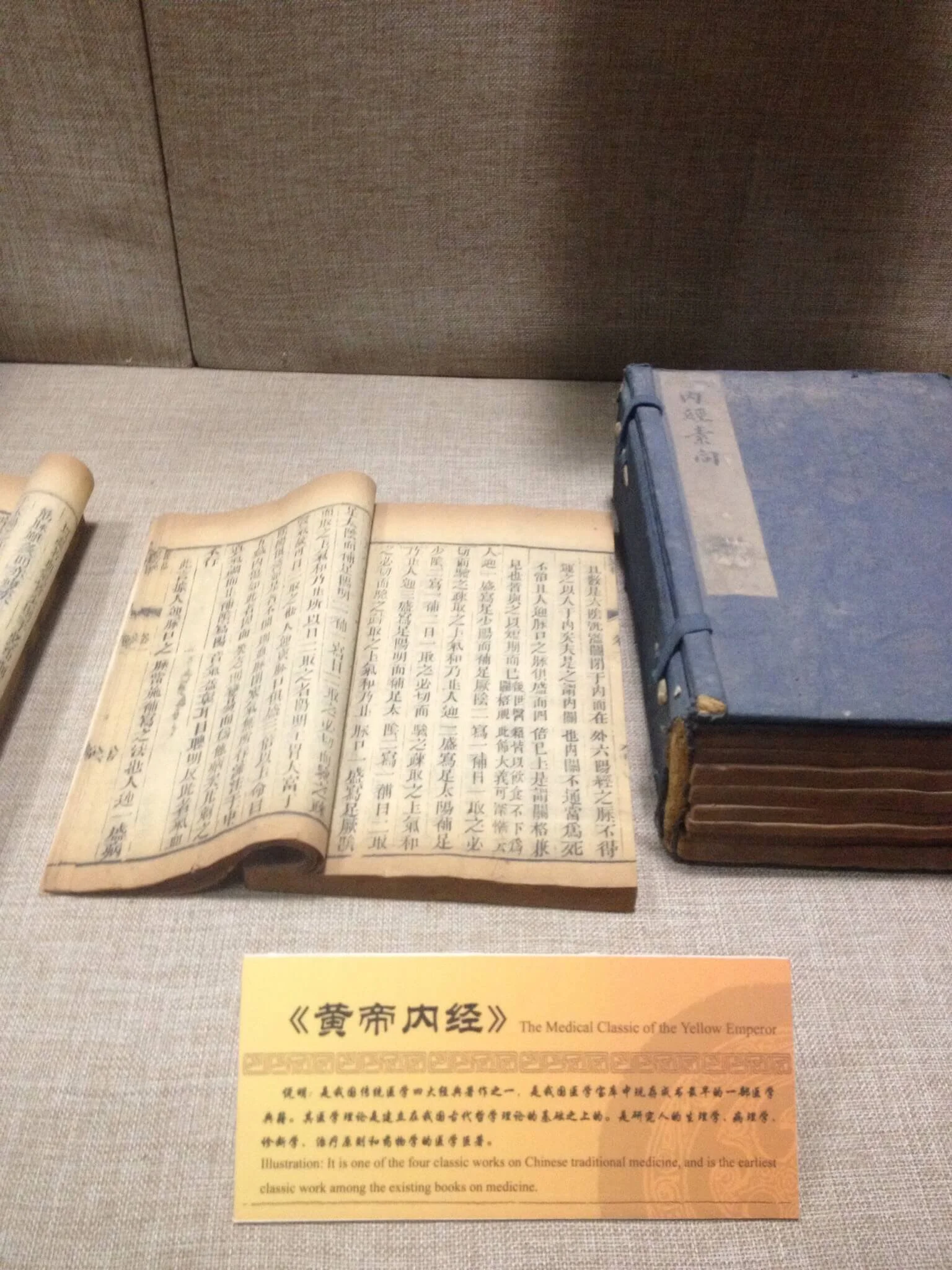 The image shows an open ancient Chinese medical book with traditional Chinese characters, placed on a table against a brown textured background. There are also other closed books nearby. A yellow informational card with Chinese and English text descr