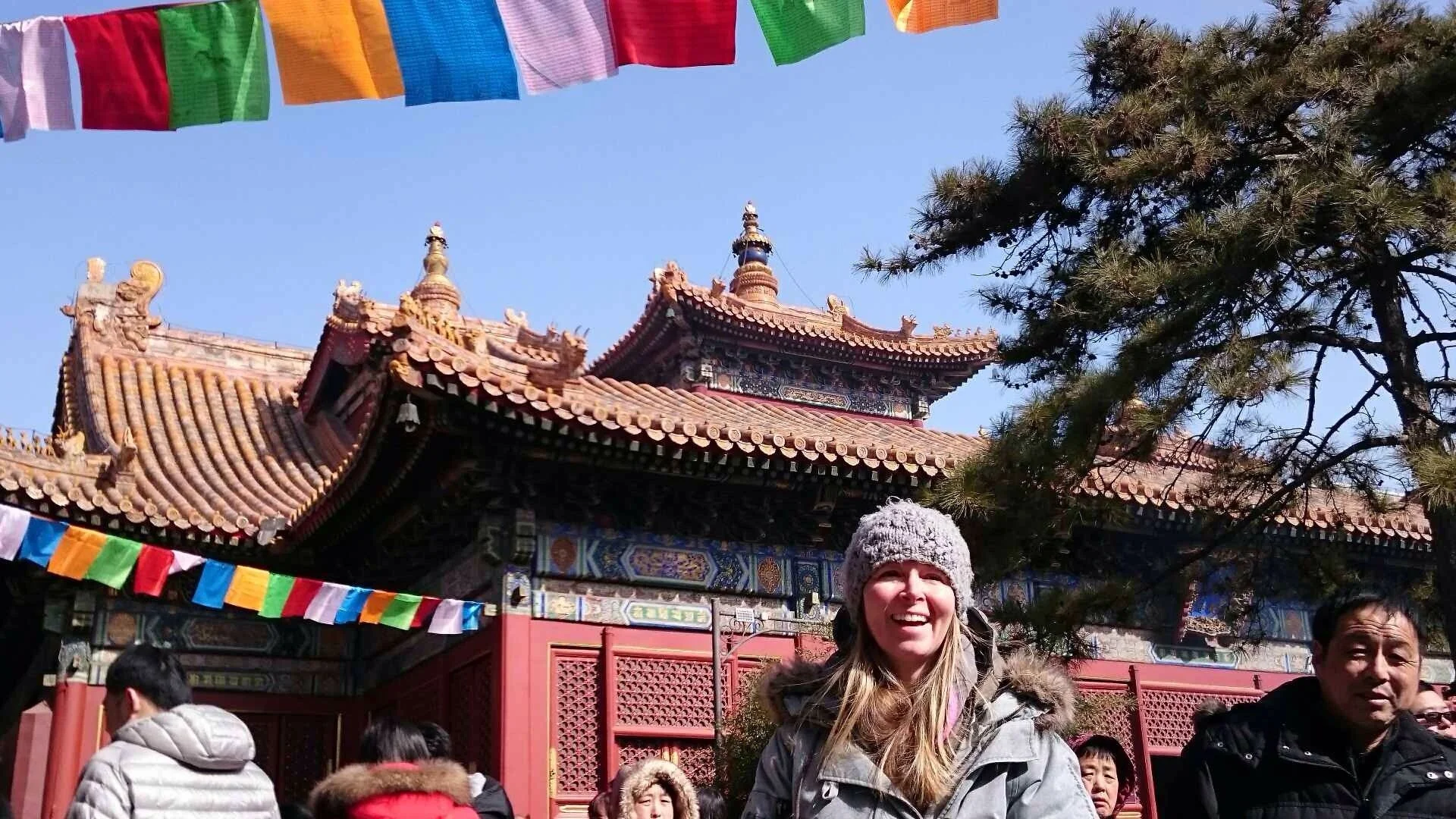 A woman with a gray hat smiling in front of a traditional Asian temple with red and gold roofs, decorated with colorful prayer flags, and surrounded by trees and a crowd of people.