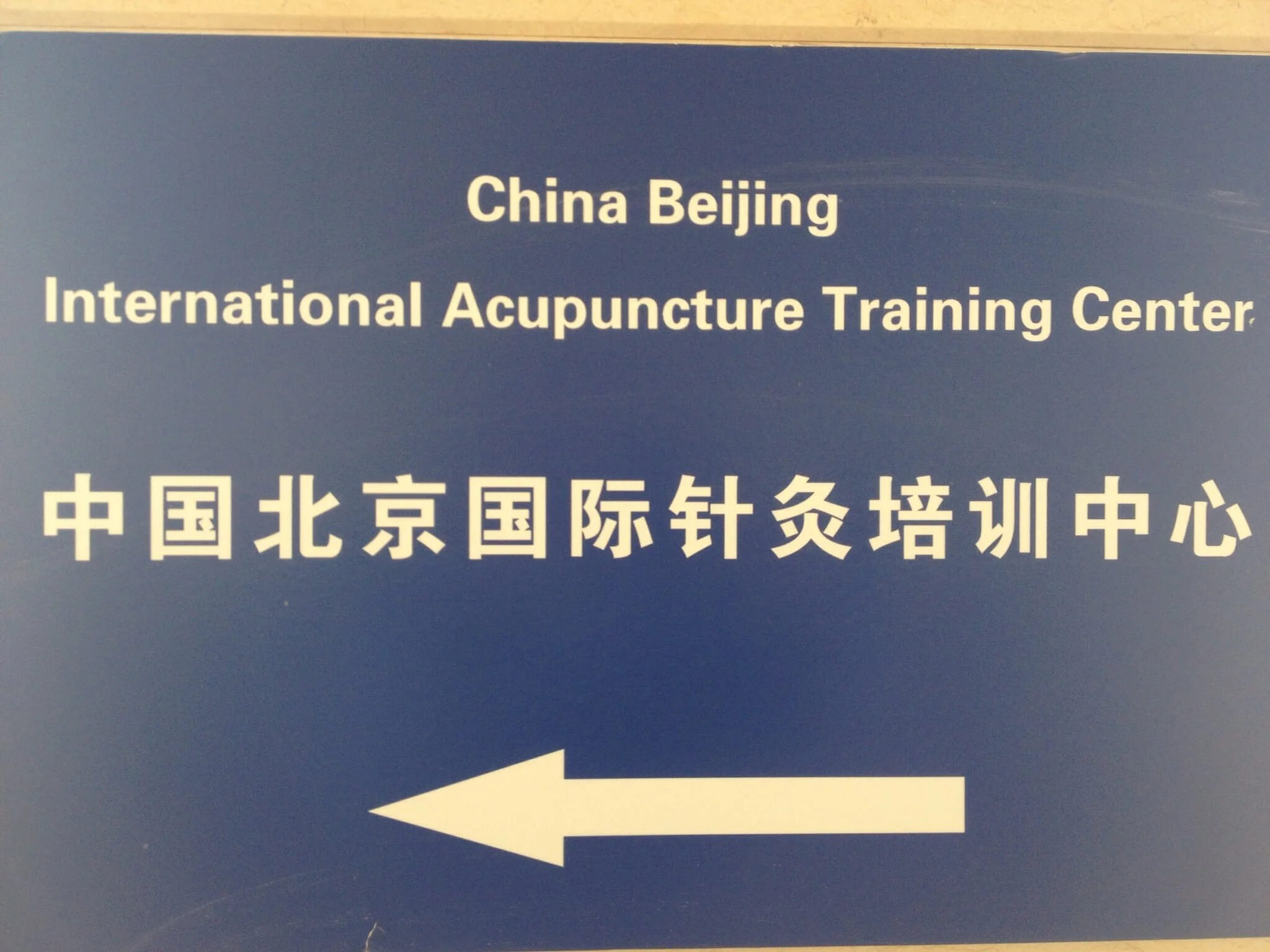 Sign with white arrow pointing left on a blue background, featuring Chinese characters and English text that reads 'China Beijing International Acupuncture Training Center'.
