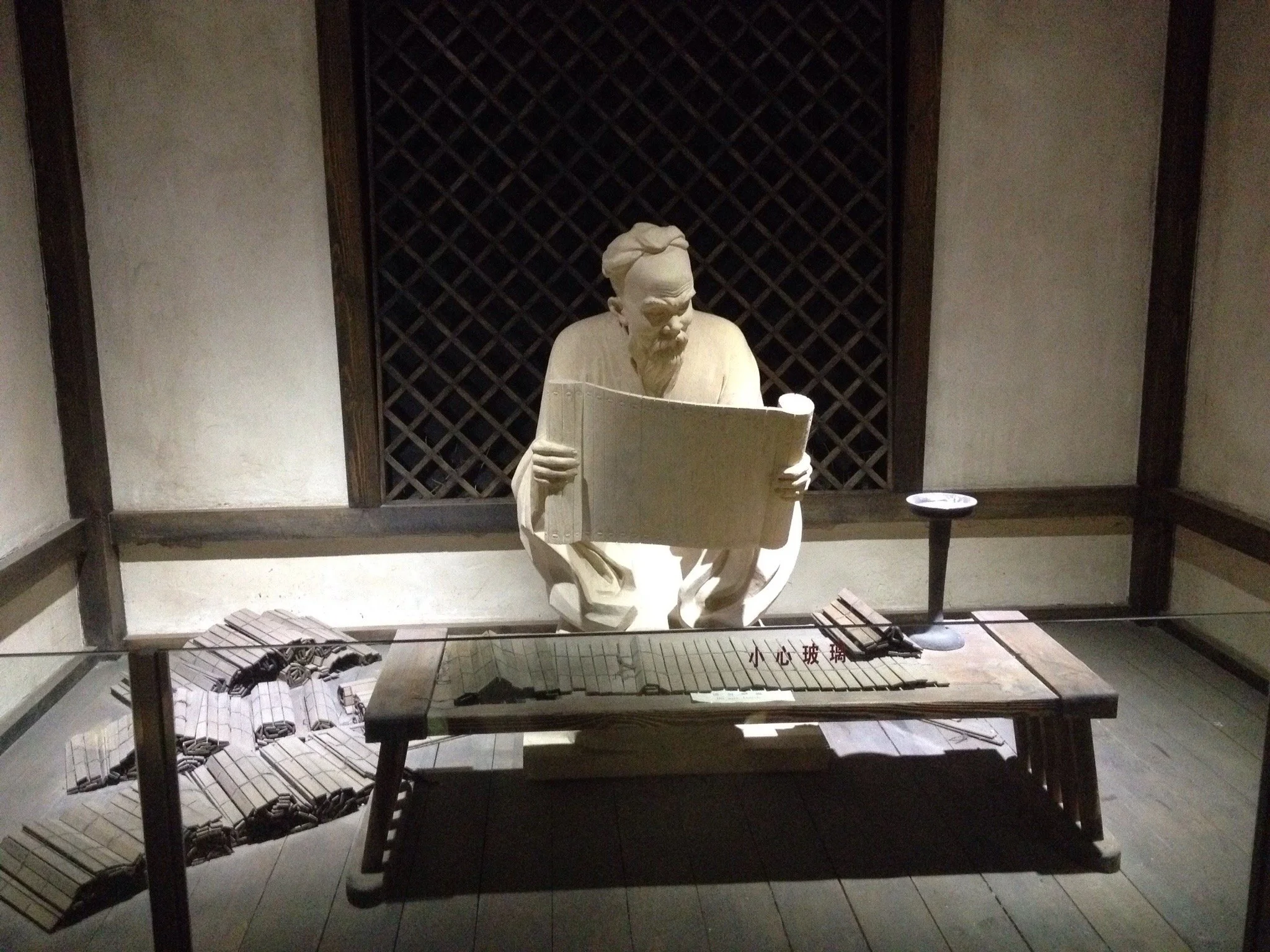 A white sculpture of a man with a beard and traditional clothing, seated and reading a book. The sculpture is displayed behind a glass case in a museum setting, with a dark wooden lattice background and a small table with model architectural structur