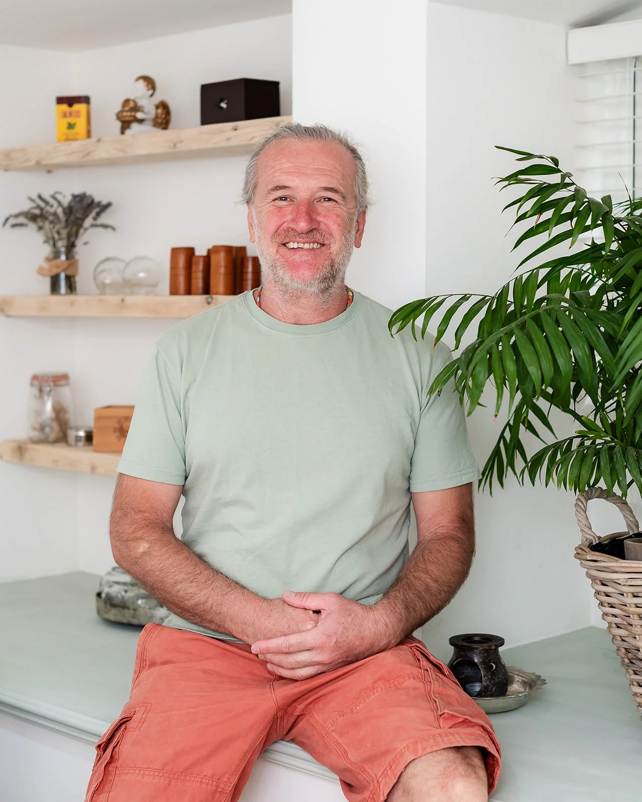 A smiling middle-aged man with gray hair and beard, wearing a light green t-shirt and orange shorts, sitting on a bench in a minimalist room with wooden shelves and green potted plant.