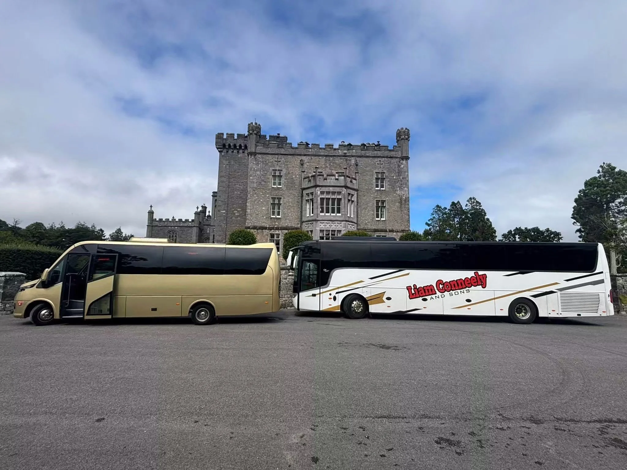 Two tour buses parked in front of a castle, with trees and a cloudy sky in the background.