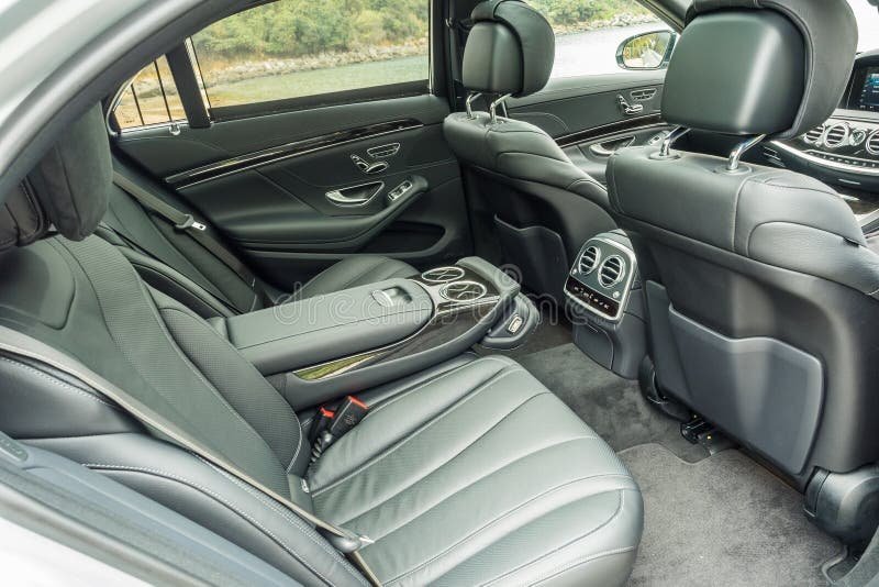 The interior of a luxury car showing black leather seats, back of the front seats with built-in screens, rear air vents, and control panel, with a view of the front dashboard and windows.