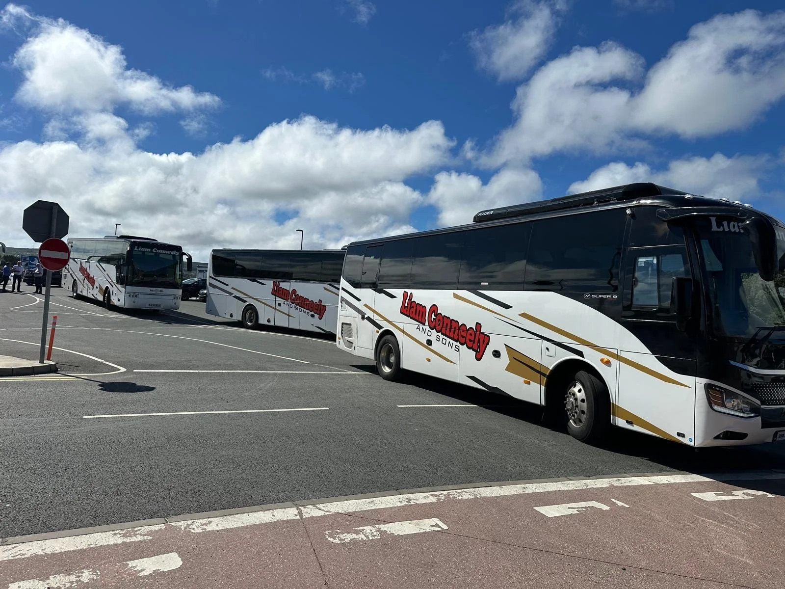 Several white buses with black and gold accents, parked in a lot under a partly cloudy sky, with some people standing nearby. The buses are labeled 'Liam Connelly and Sons.'