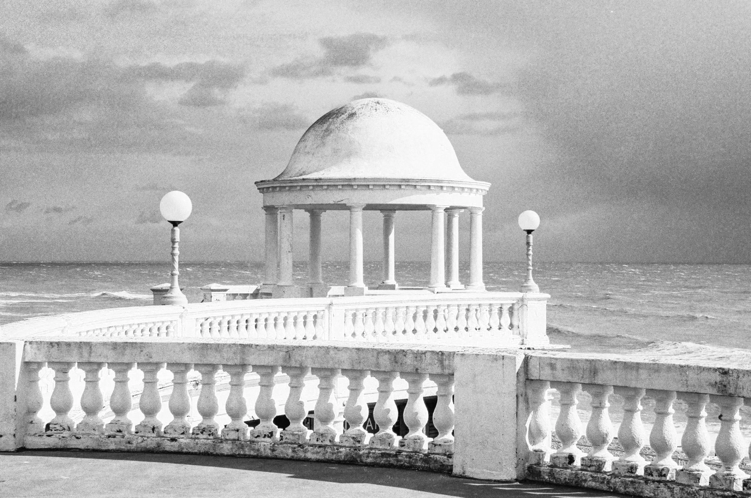 Black and white photograph of a seaside pavilion with a domed roof supported by columns, surrounded by a decorative balustrade, and flanked by two street lamps, with clouds and the ocean in the background.