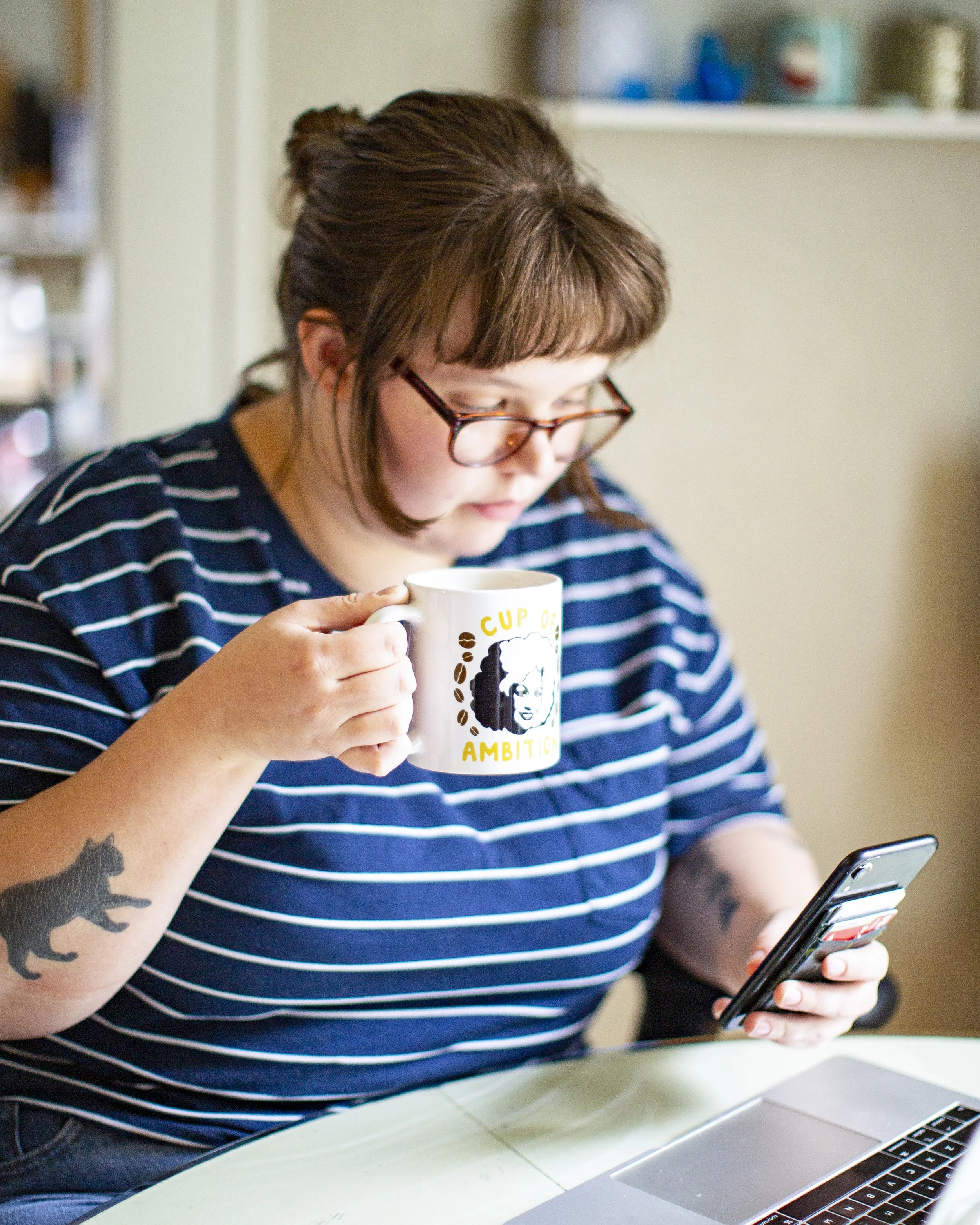Photo of Marin looking at her phone and holding a Dolly Parton mug that says, "Cup of Ambition"