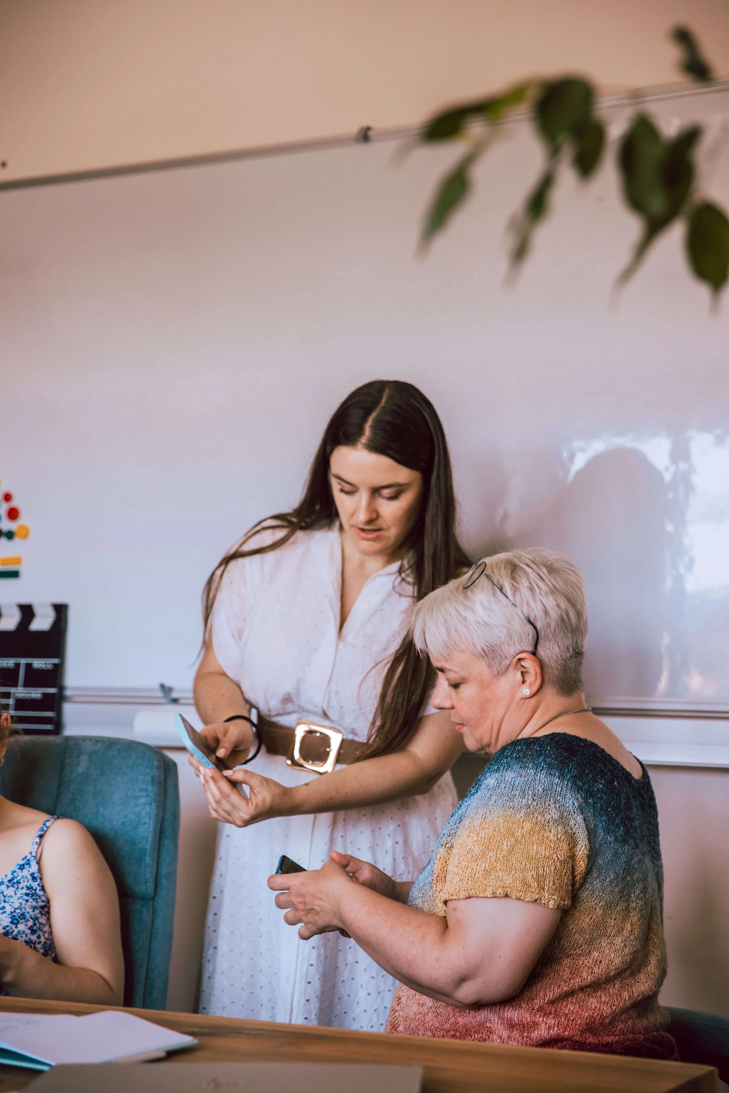 Drei Frauen, die in einem Raum an einem Tisch sitzen. Zwei Frauen sind im Gespräch und eine im Hintergrund, während eine weitere Frau im Vordergrund ein Smartphone nutzt. Im Hintergrund eine weiße Tafel und einige Büroartikel.
