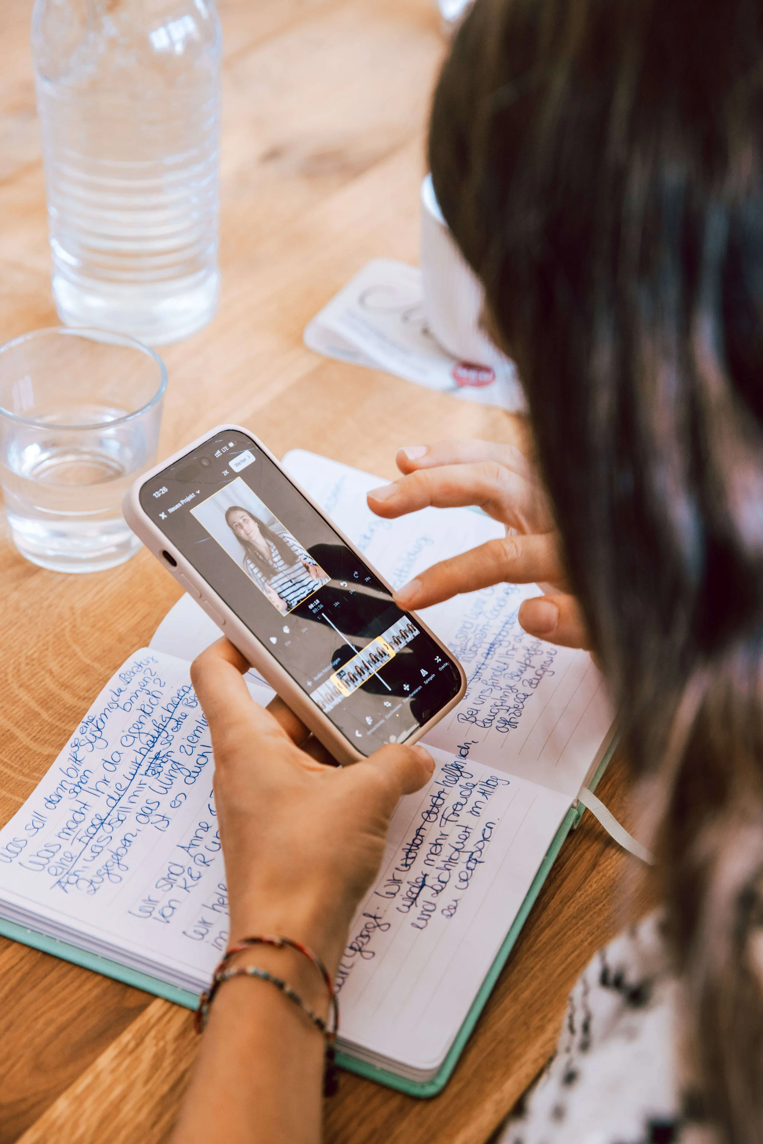 Person filmt eine junge Frau mit einem Smartphone während sie in einem Notizbuch schreibt, auf einem Holztisch stehen Wasserflaschen und Gläser.