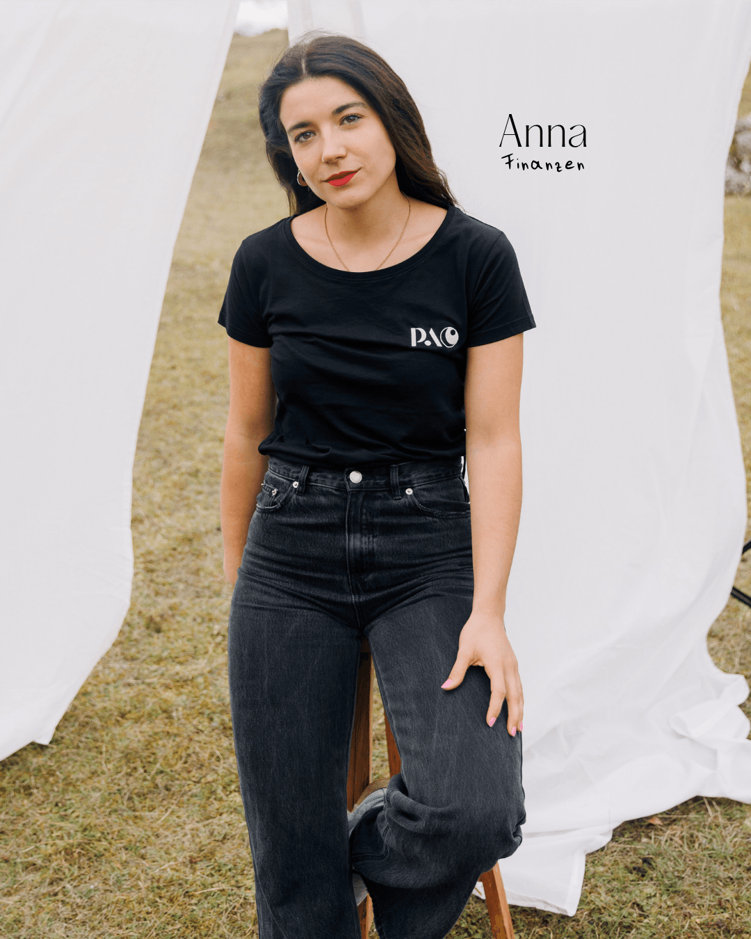 Eine junge Frau mit dunklen Haaren, rotem Lippenstift und schwarzen Jeans sitzt vor einem weißen Hintergrund im Freien. Sie trägt ein schwarzes T-Shirt mit weißem Logo. Handschriftlich steht neben ihr: "Anna" und "Finanzen".