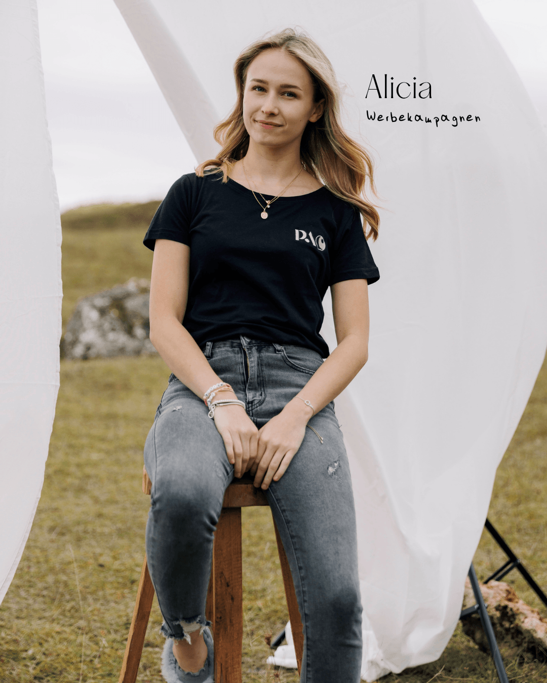 Junge Frau sitzt auf einem Holzstuhl vor weißen Stoffbahnen, trägt schwarze T-Shirt mit weißem Logo, graue Jeans mit Rissen, trug Schmuck, im Freien.