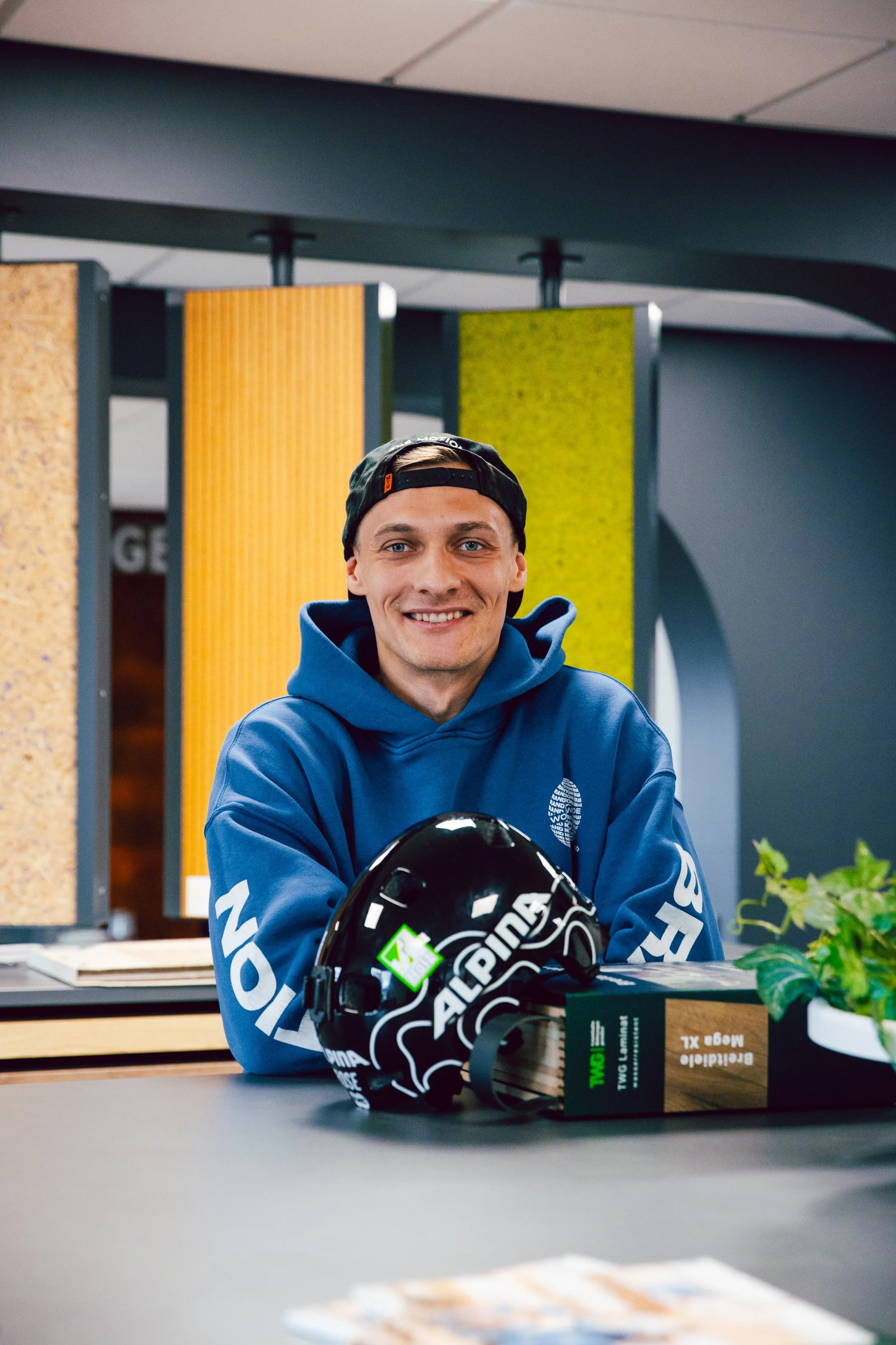 Ein junger Mann in blauer Kleidung sitzt an einem Tisch in einem Raum, mit einem Helm vor sich, auf dem er lächelt. Im Hintergrund sind bunte vertikale Paneele an der Wand.