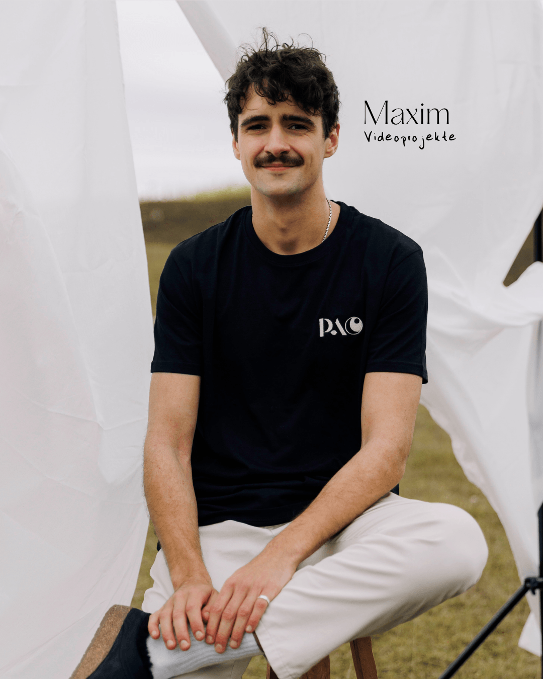 Junger Mann sitzt draußen vor weißem Hintergrund mit Text 'Maxim Videoprojekte' und trägt ein schwarzes T-Shirt mit 'PAC'-Logo.