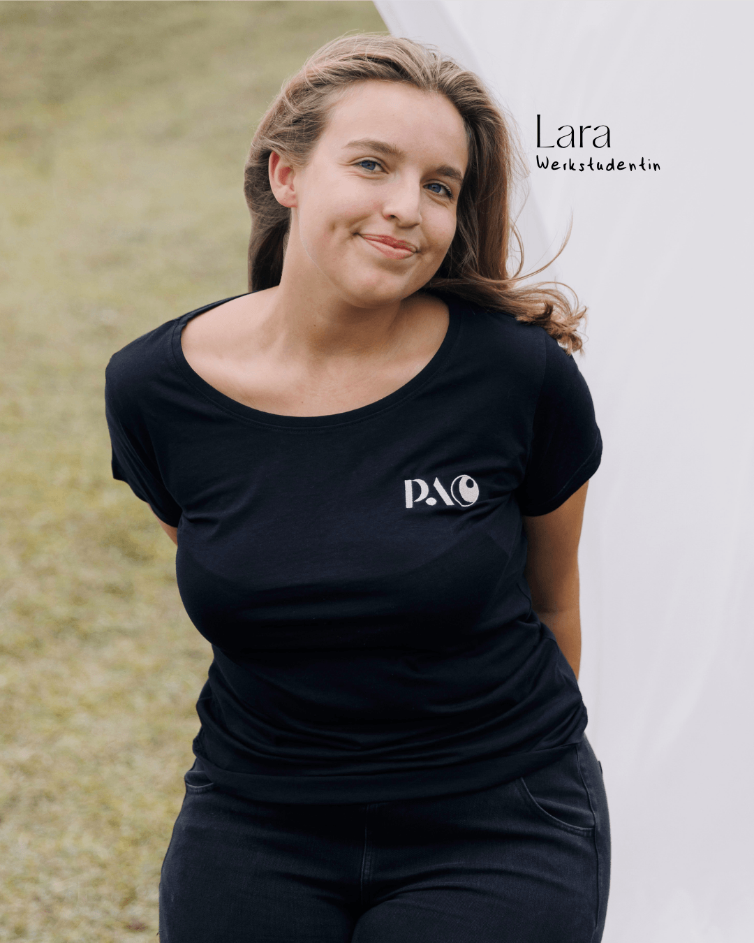 Junge Frau mit braunen Haaren, schwarzes T-Shirt und dunklen Hosen steht im Freien, lächelt und posiert.