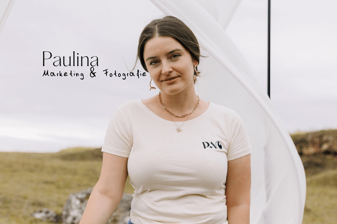 Porträt einer jungen Frau in einem weißen T-Shirt und blauen Jeans, sitzend auf einem Holzstuhl im Freien, mit Fotografie-Equipment im Hintergrund.