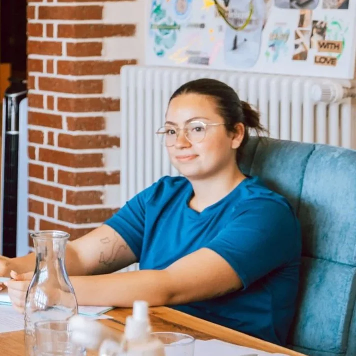 Eine junge Frau mit Brille und blauer T-Shirt sitzt an einem Tisch in einem Raum mit roten Backsteinwänden und einer Pinnwand im Hintergrund.