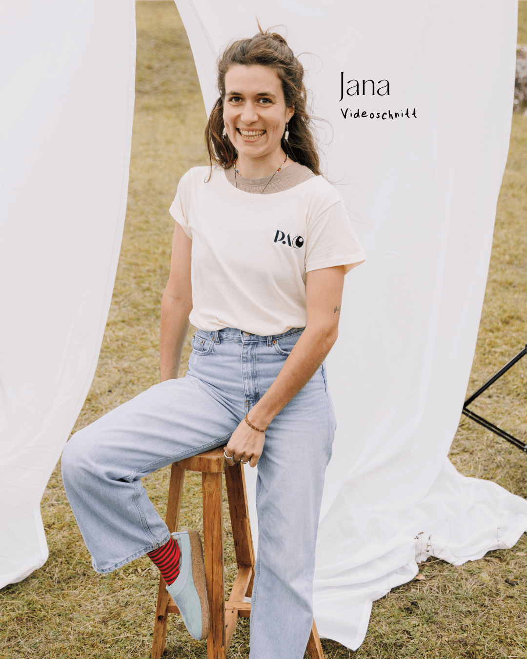 Junge Frau sitzt auf einem Schemel, lächelt, trägt weißes T-Shirt, Jeans und bunte Socken, im Freien vor weißem Hintergrund, Text auf der Bild: Jana, Videoschnitt.