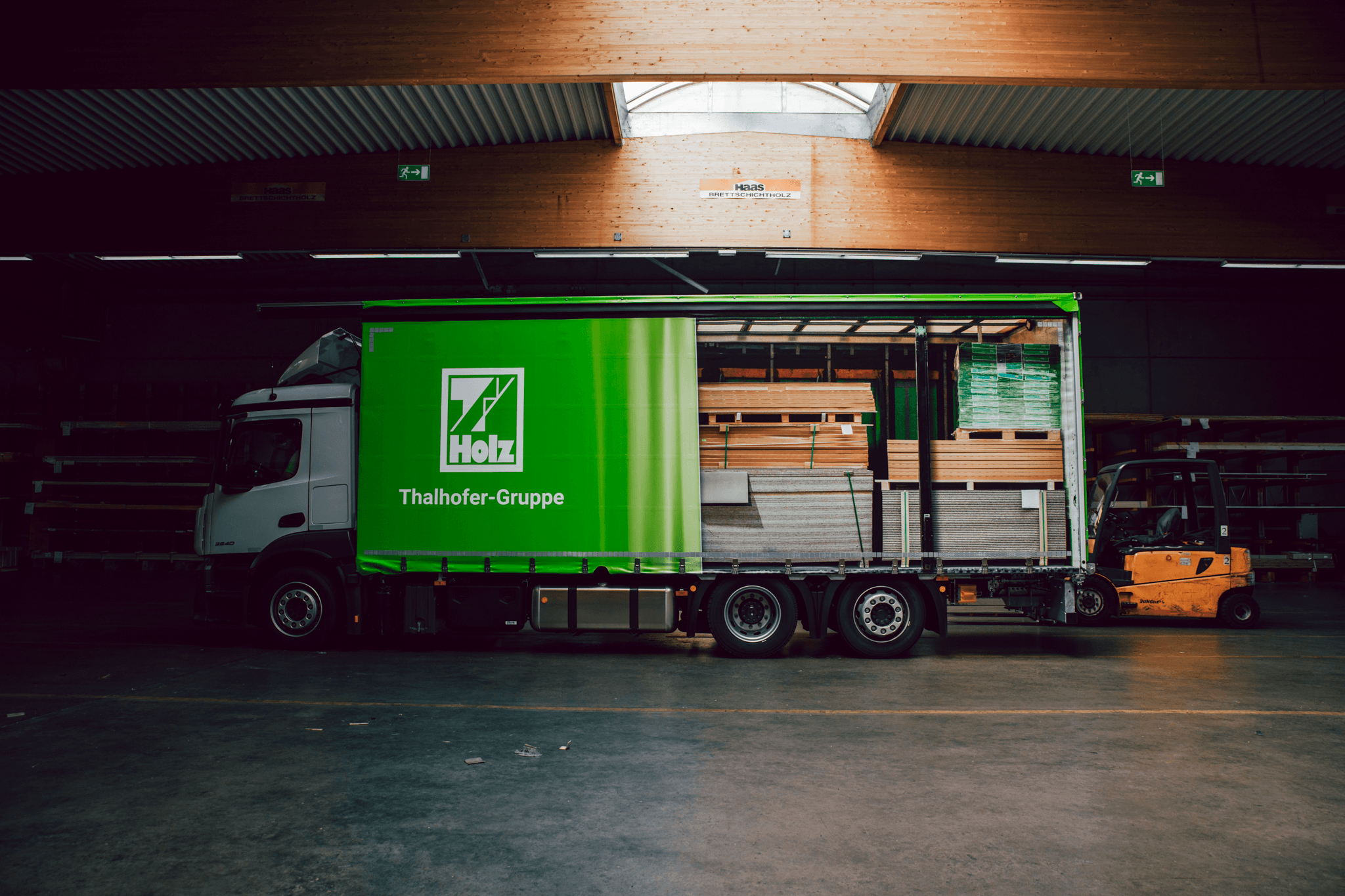 Lagerraum mit grünem Lieferwagen, der Holz und Baummaterialien transportiert, im Holzwerk Thalhofer-Gruppe in einer industriellen Umgebung.