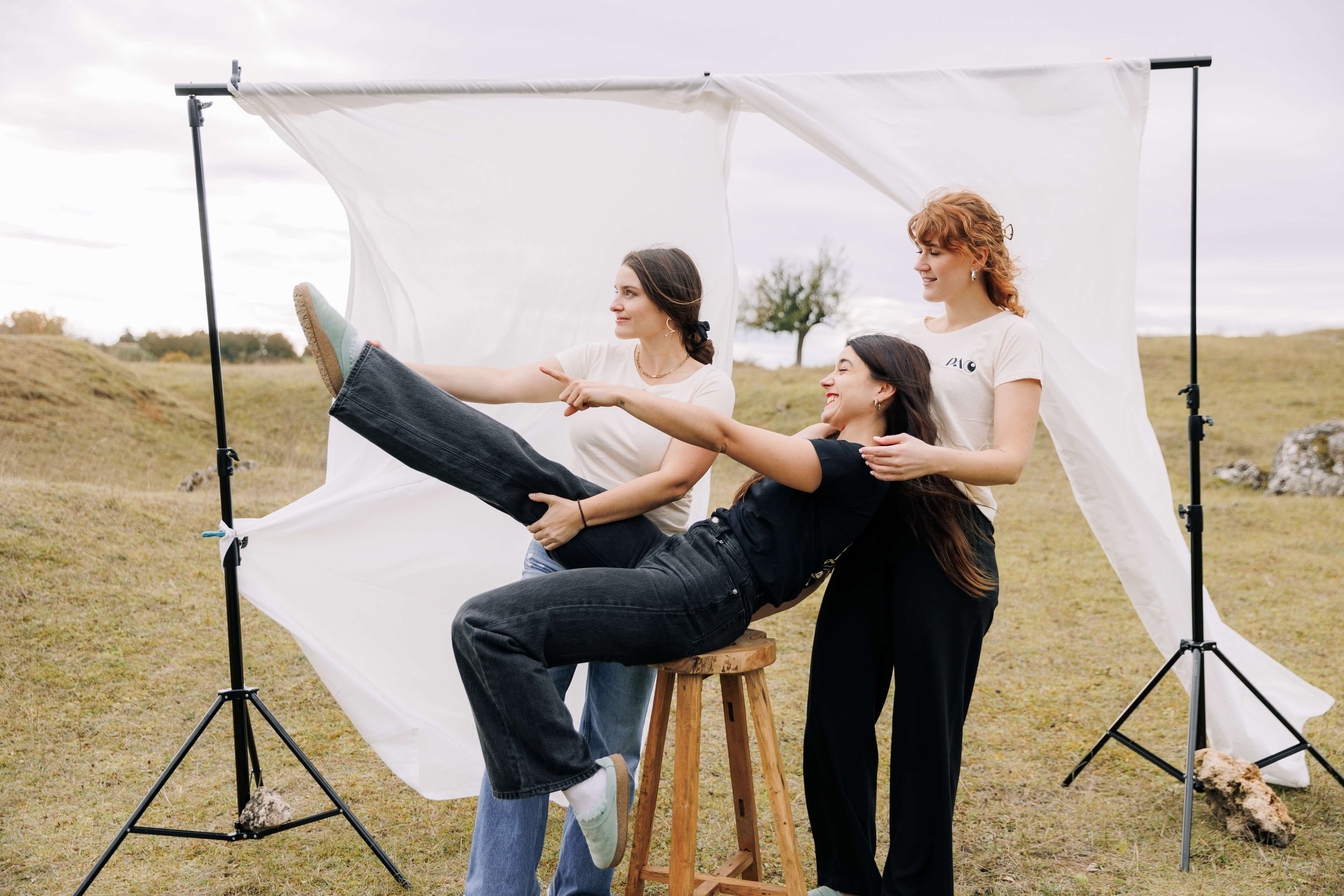 Drei Frauen, die bei einem Fotoshooting im Freien auf einer Wiese stehen, eine der Frauen sitzt auf einem Stuhl und wird von den anderen beiden gehalten. Es ist bewölkt, und ein weißer Hintergrundschirm ist sichtbar.