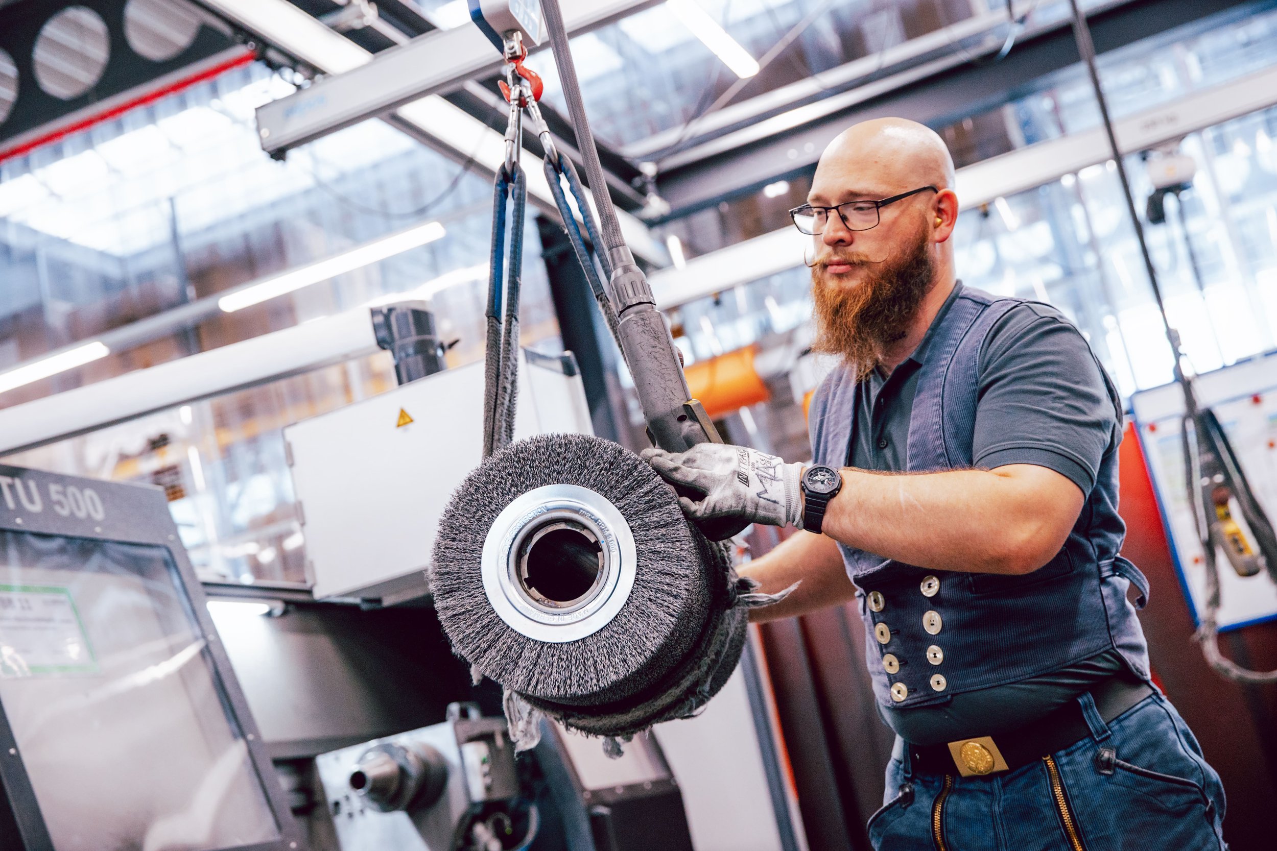 Mann mit Brille und Bart arbeitet in einer Werkstatt mit Schleifmaschine, trägt schwarze Handschuhe, dunkles T-Shirt und Jeans, hinter ihm sind Maschinen und Regale sichtbar.