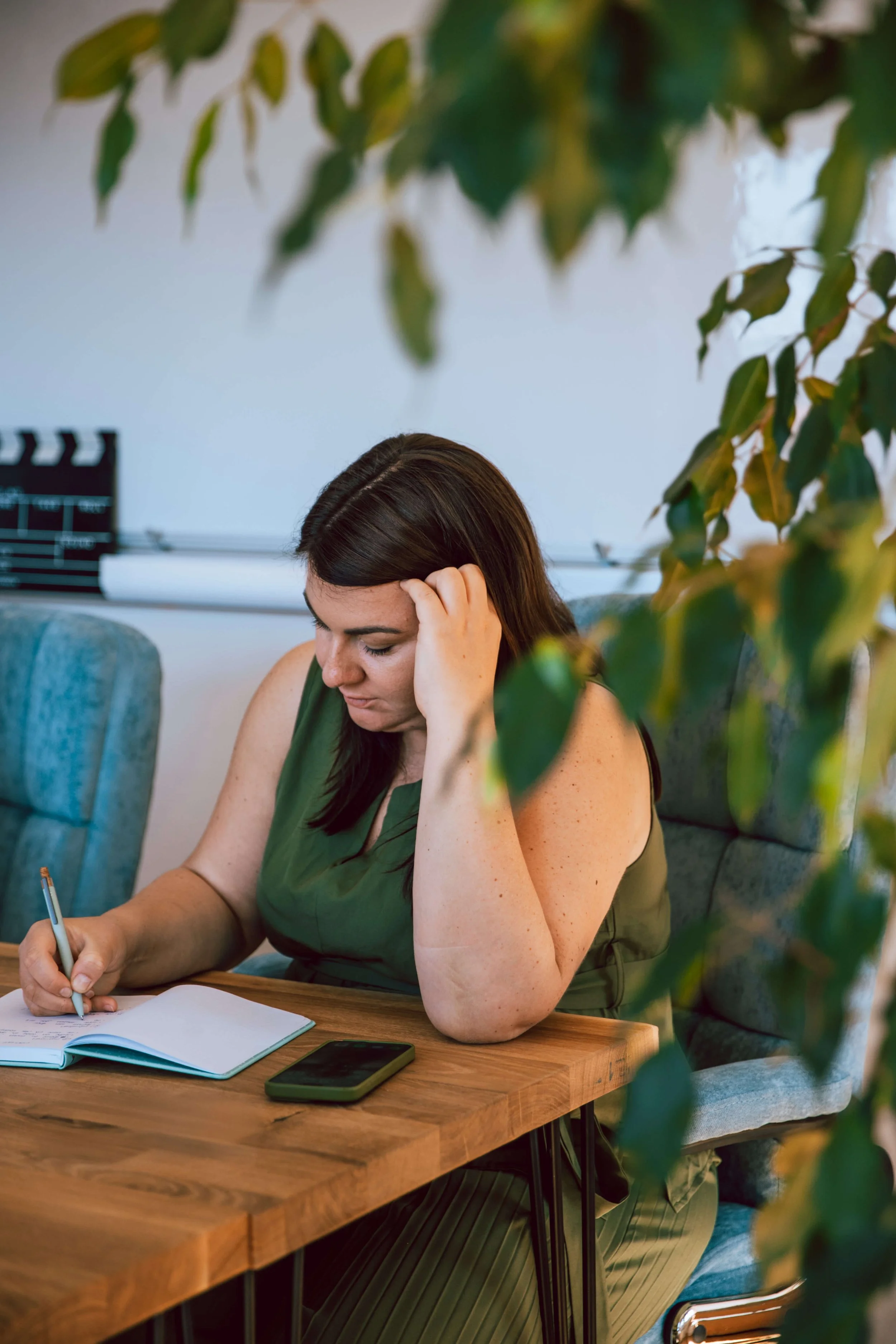 Eine Frau mit braunen Haaren, die an einem Holztisch sitzt, einen Stift hält, in ein Notizbuch schreibt und auf ihrem Handy schaut, während sie nachdenklich ist, im Hintergrund ein künstlerischer Raum mit einem Filmklappe und einer weißen Wand.