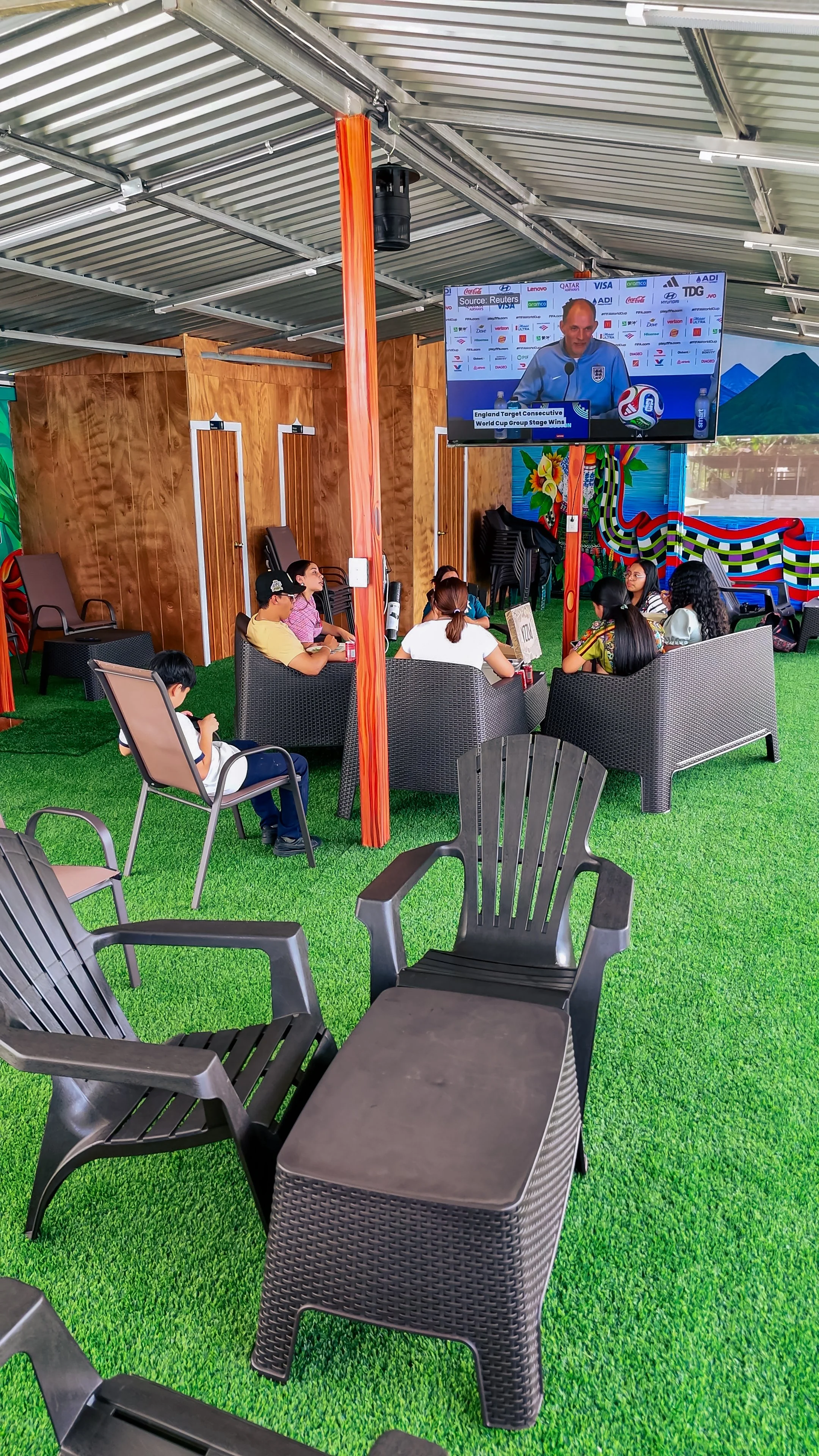 People sitting and watching a large television screen outdoors under a metal roof. The scene appears to be a viewing area with chairs on artificial green grass, with colorful decorative elements on the walls.