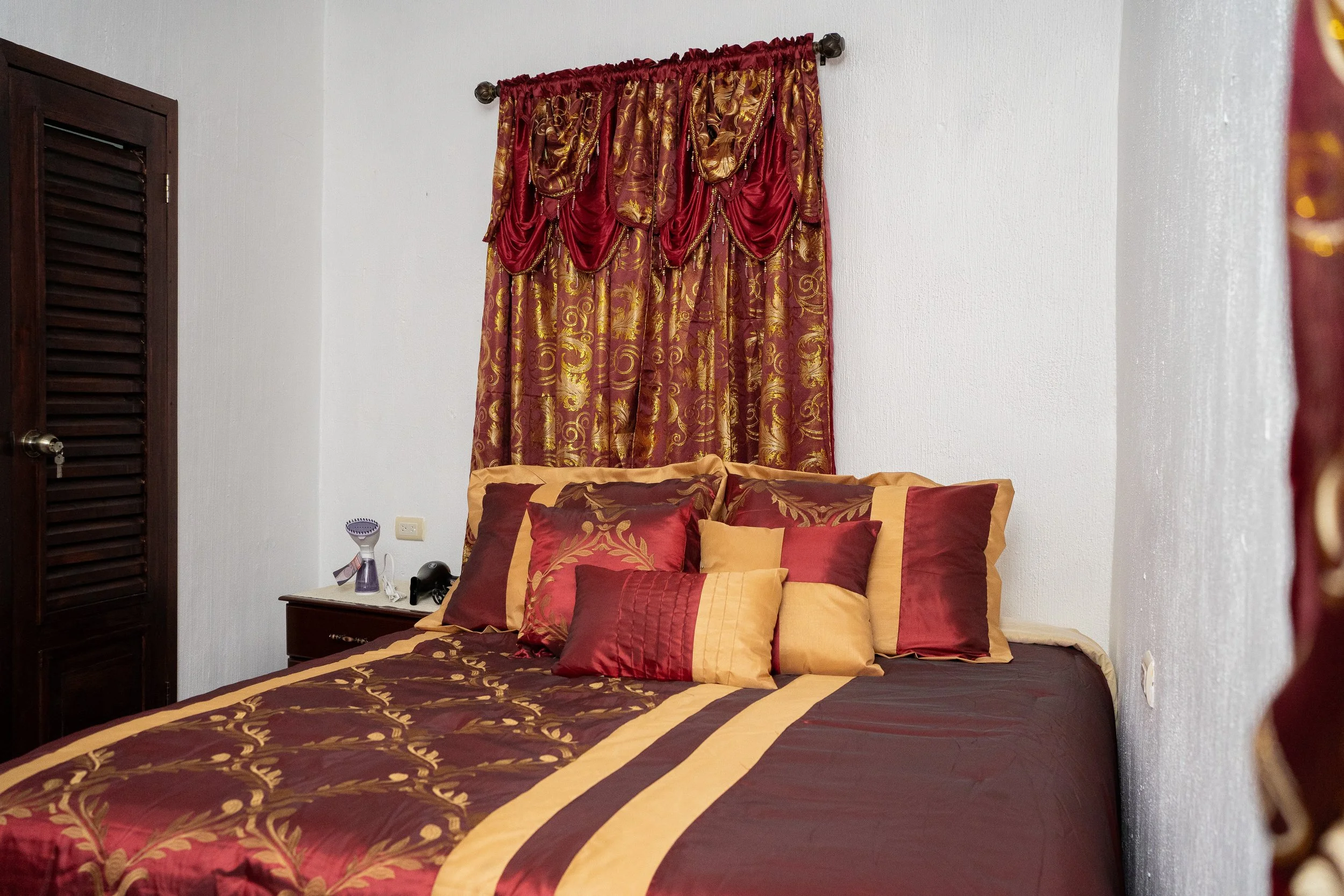 A bedroom with a bed covered in a maroon and gold bedspread, surrounded by matching pillows. Behind the bed is a decorative curtain with red, gold, and maroon fabric, gathered at the top and adorned with tassels. To the left, there is a wooden closet
