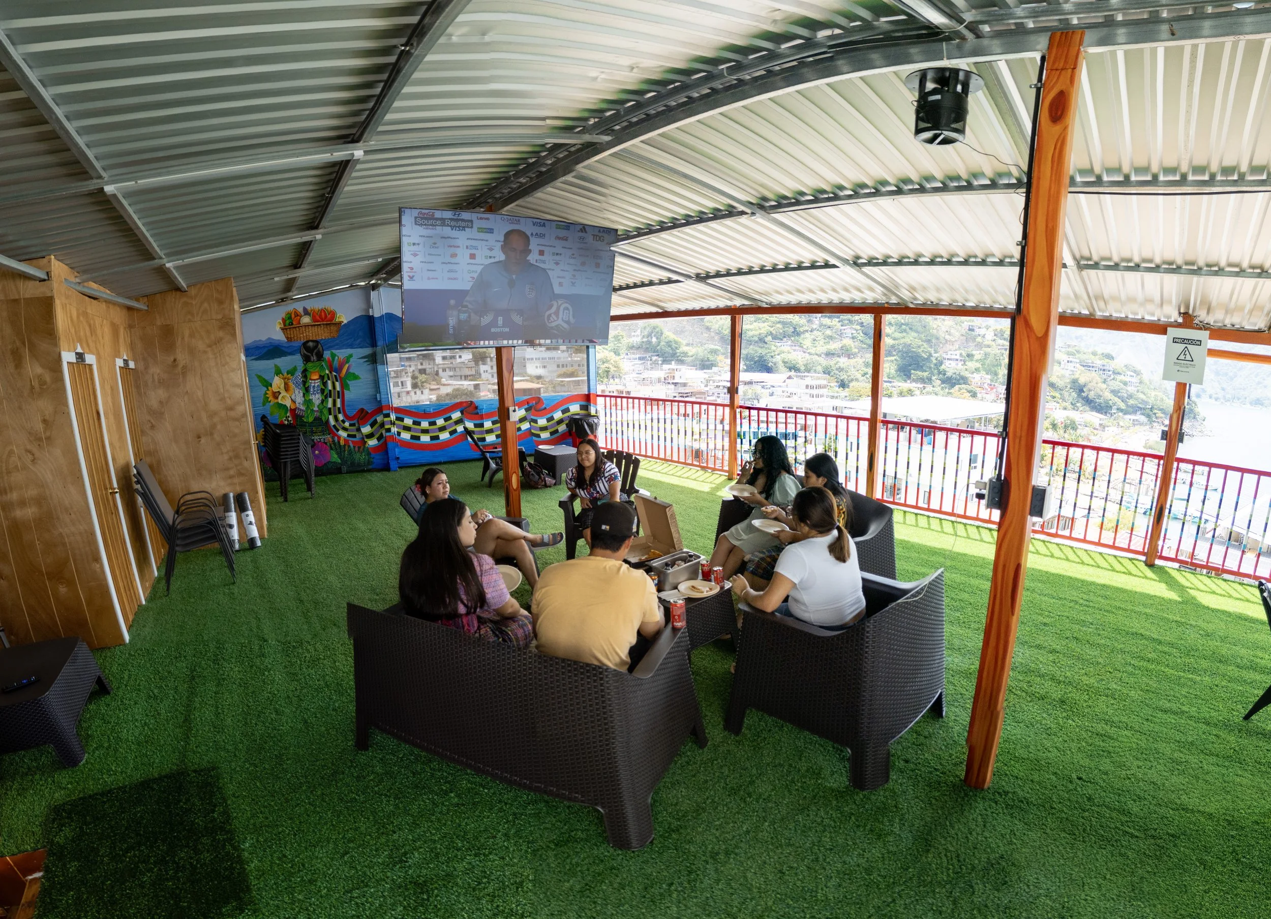 People seated at tables on a green outdoor terrace overlooking a cityscape, with a large screen showing a man speaking at a press conference in the background.