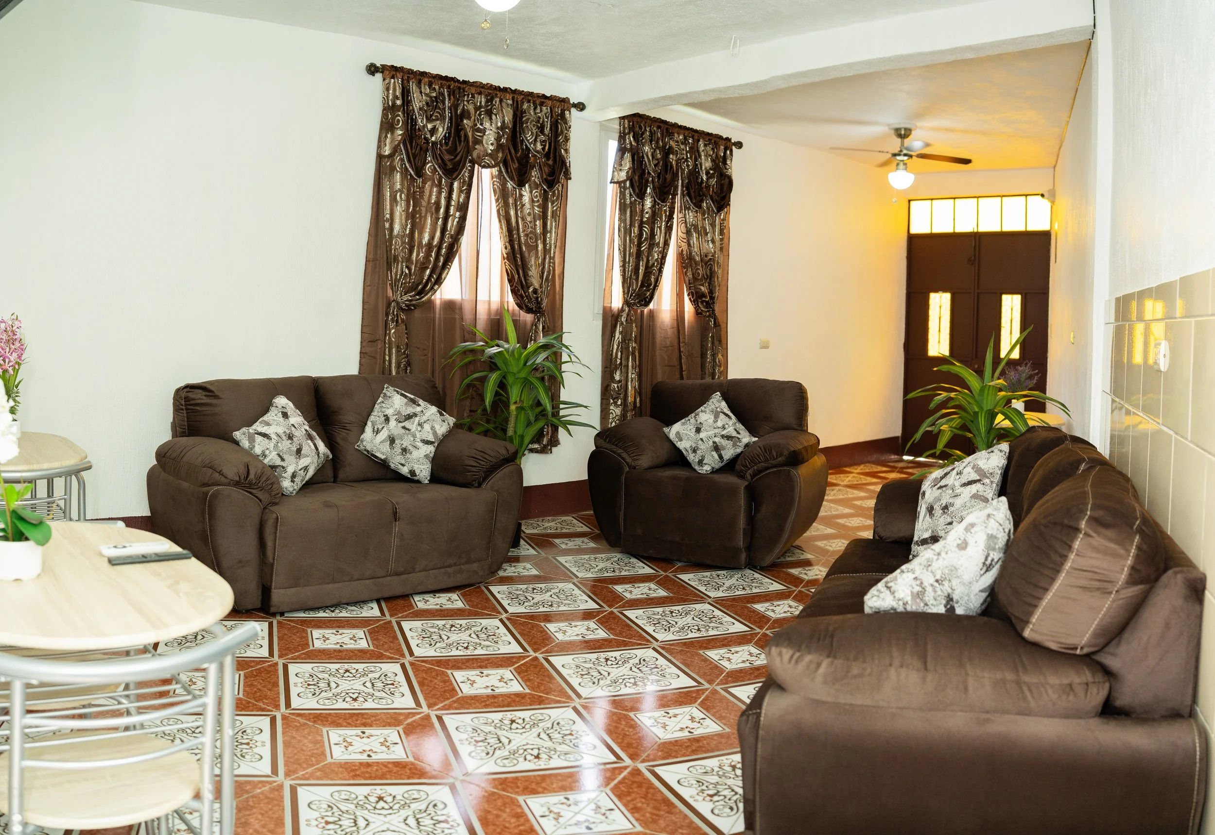 Living room with brown sofas, patterned cushions, green plants, large windows with ornate brown curtains, ceiling fan, and tiled floor.