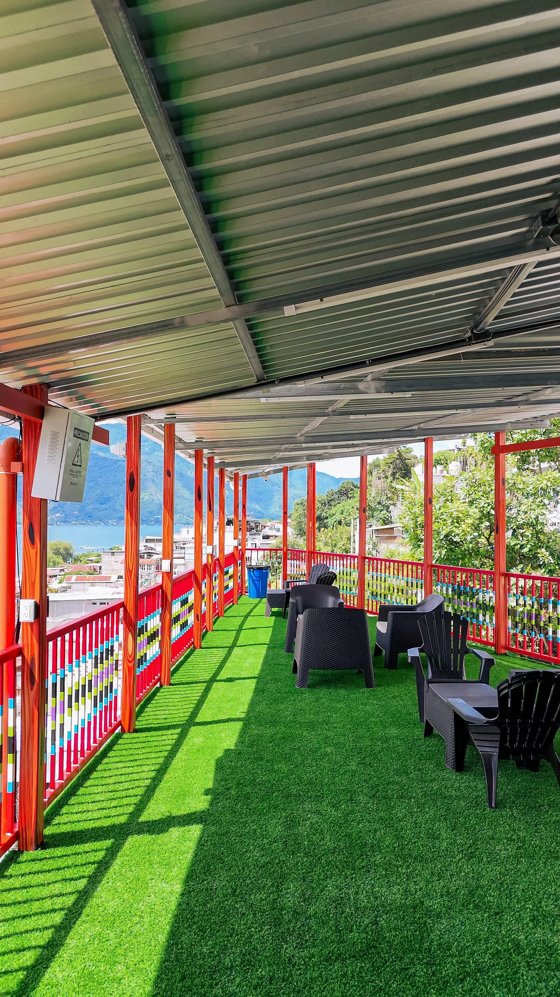 An outdoor rooftop patio with colorful railing, black chairs, artificial green grass flooring, and a metal roof, with a scenic view of mountains in the background.