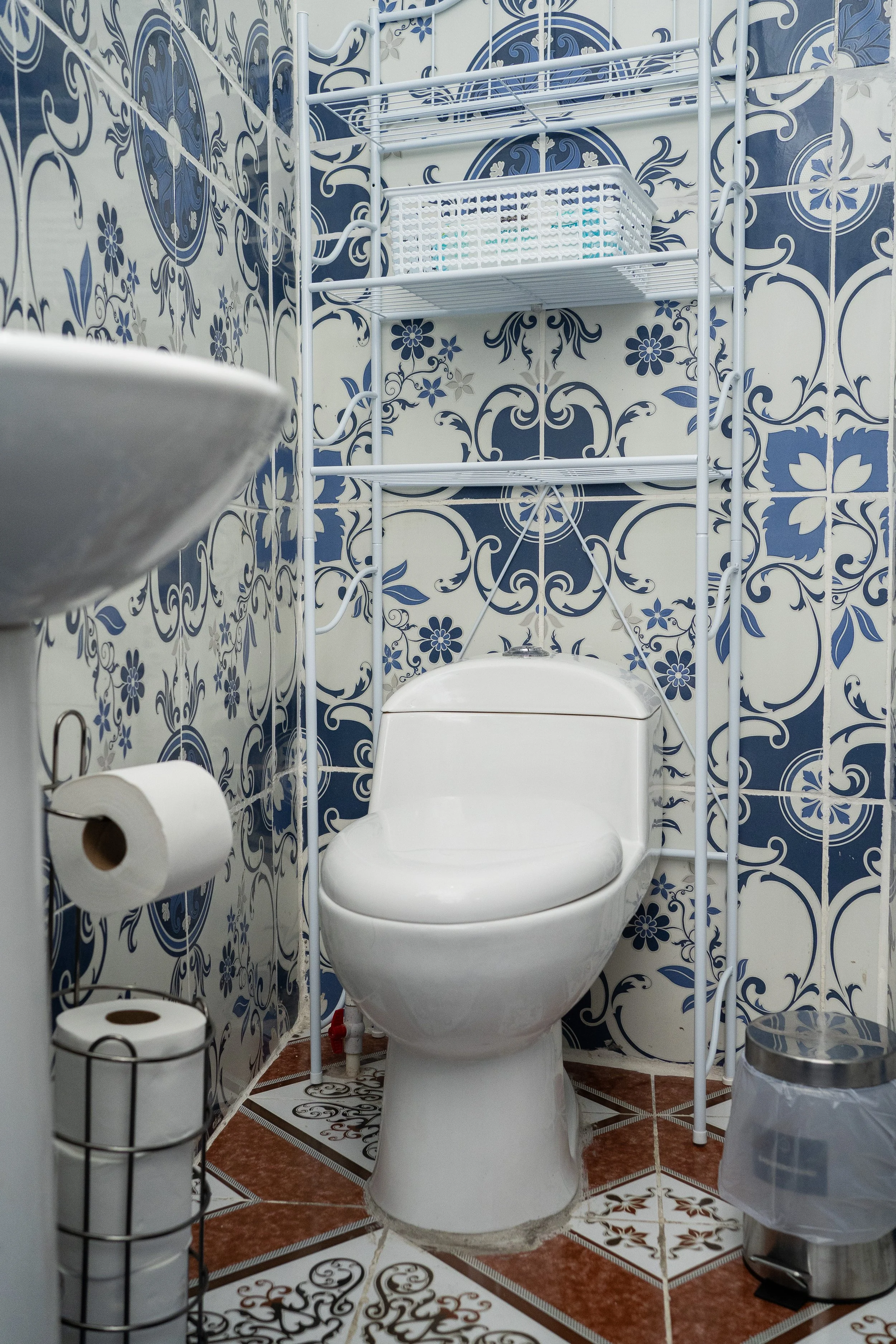 Bathroom with a white toilet behind a metal frame, surrounded by patterned blue and white tile walls, with toilet paper rolls on a stand, a trash bin, and a small shelf.