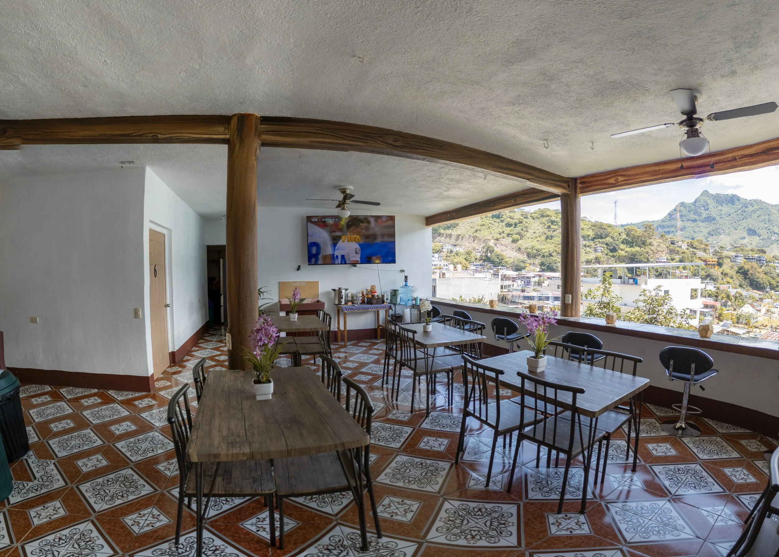 A spacious indoor area with a ceiling-mounted TV displaying a soccer game, wooden beams, ceiling fans, and large windows revealing a hilly landscape in the background. Inside, there are multiple tables with potted plants and chairs, and a kitchen cou