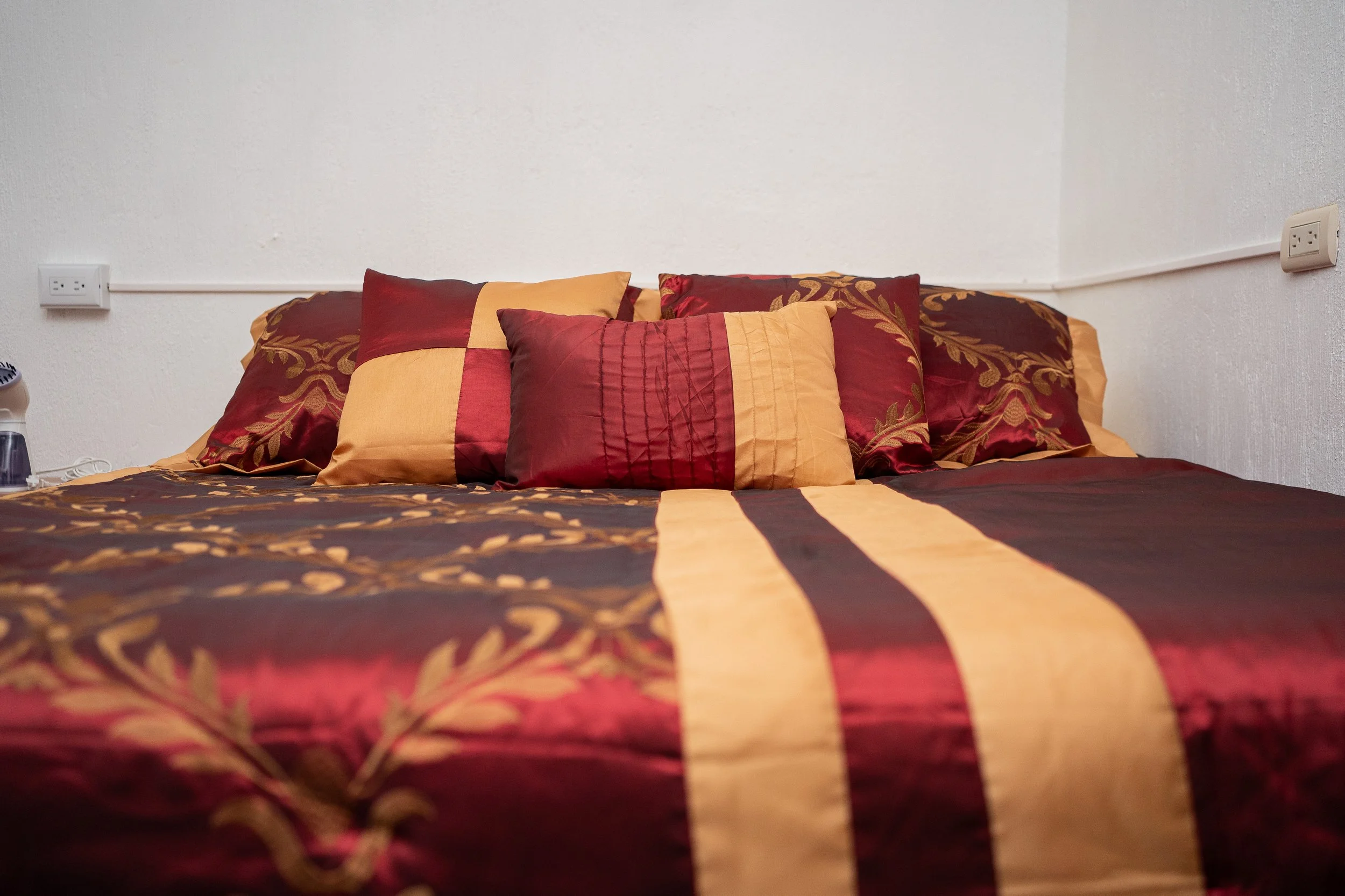 A bed with a red, orange, and gold patterned bedspread and matching pillows against a white wall with electrical outlets.
