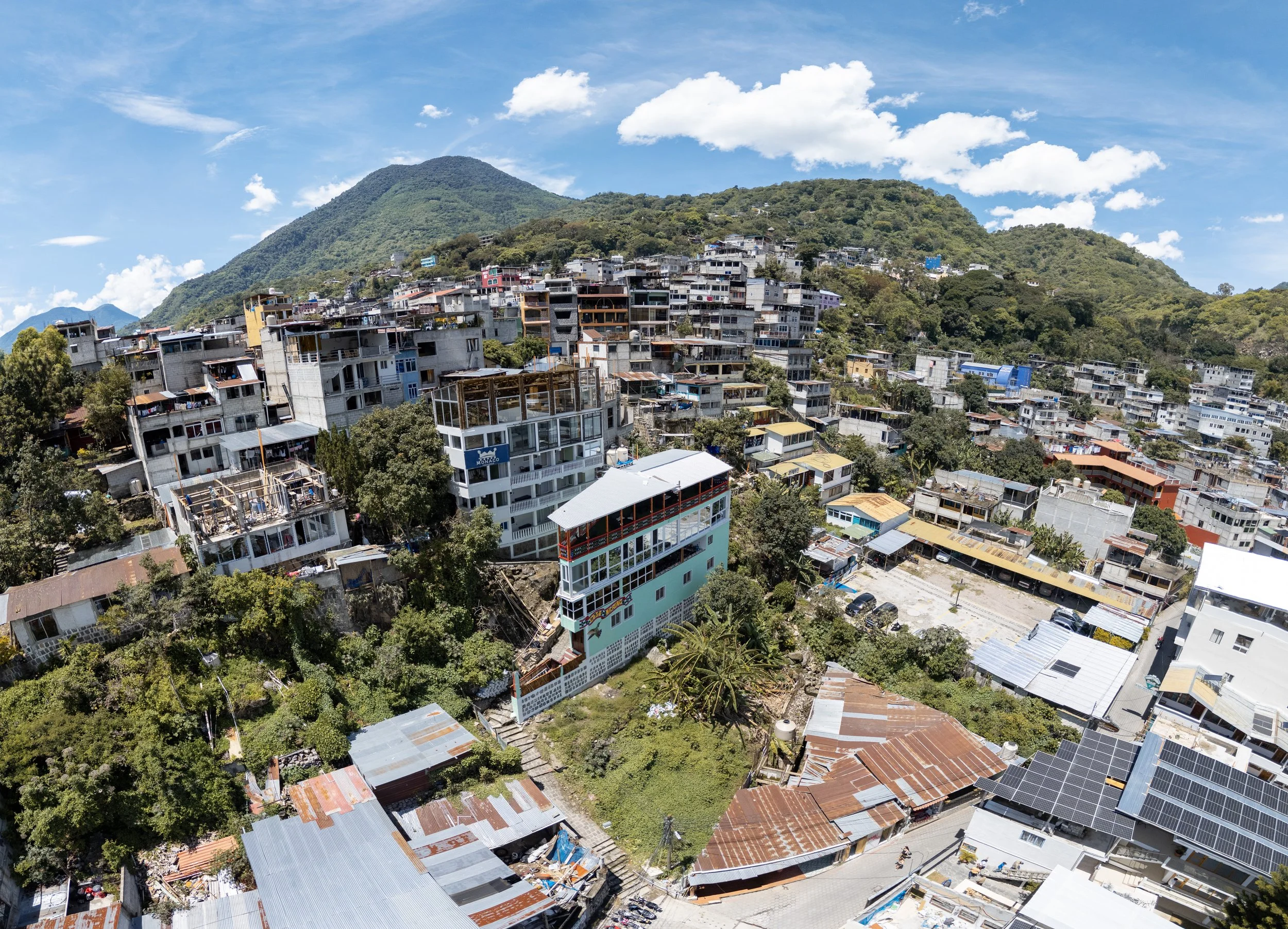 A hilly area with many residential buildings, some under construction, and lush green vegetation. The sky is partly cloudy with a few scattered white clouds.