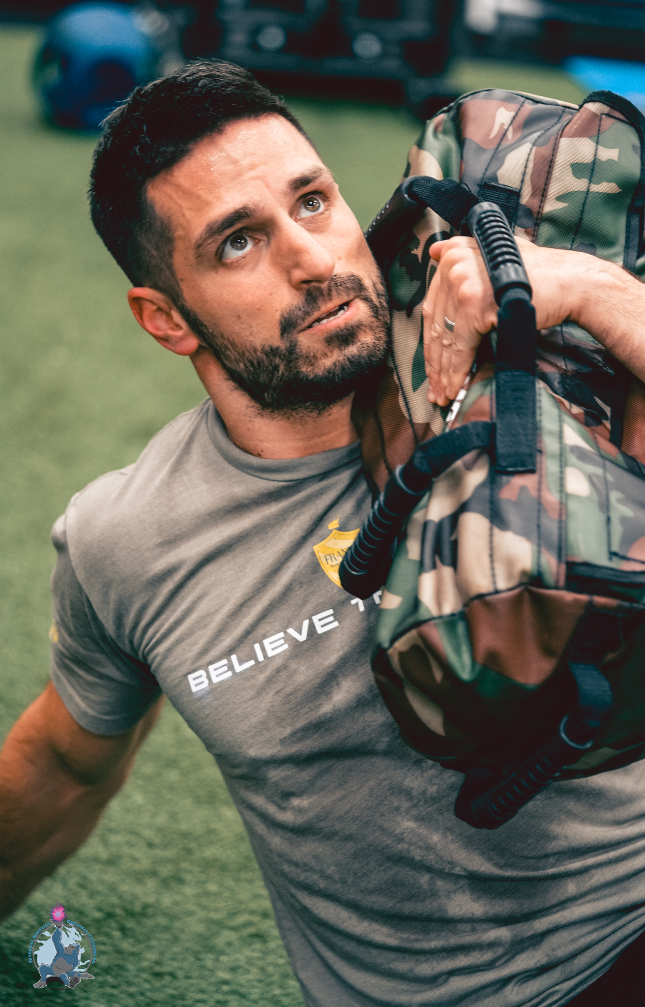 Man in gray T-shirt with 'Believe' printed on it, lying on a green turf field, holding a camouflage backpack on his shoulder, with a focused expression.
