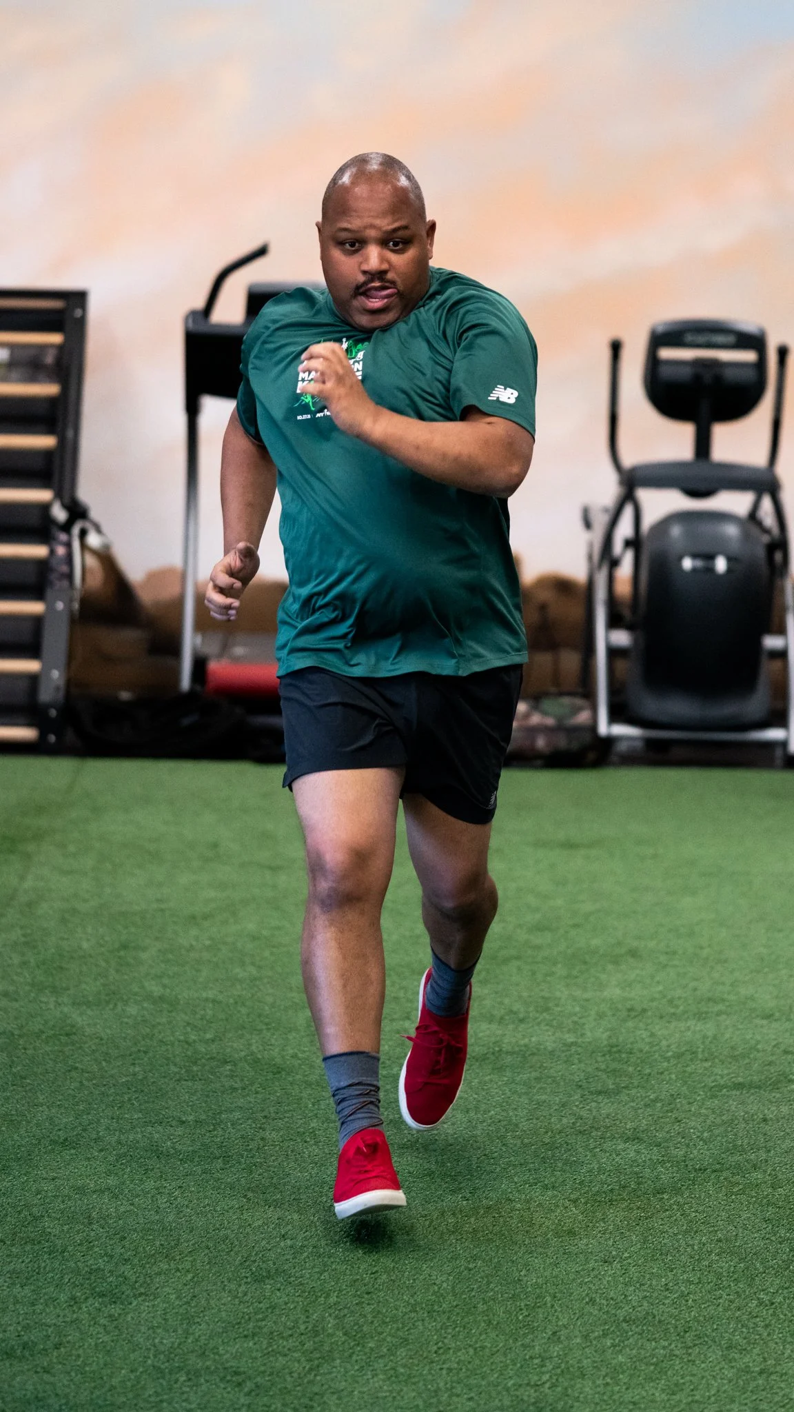 Man jogging on indoor turf wearing a green athletic shirt, black shorts, red sneakers, and gray socks with red accents.