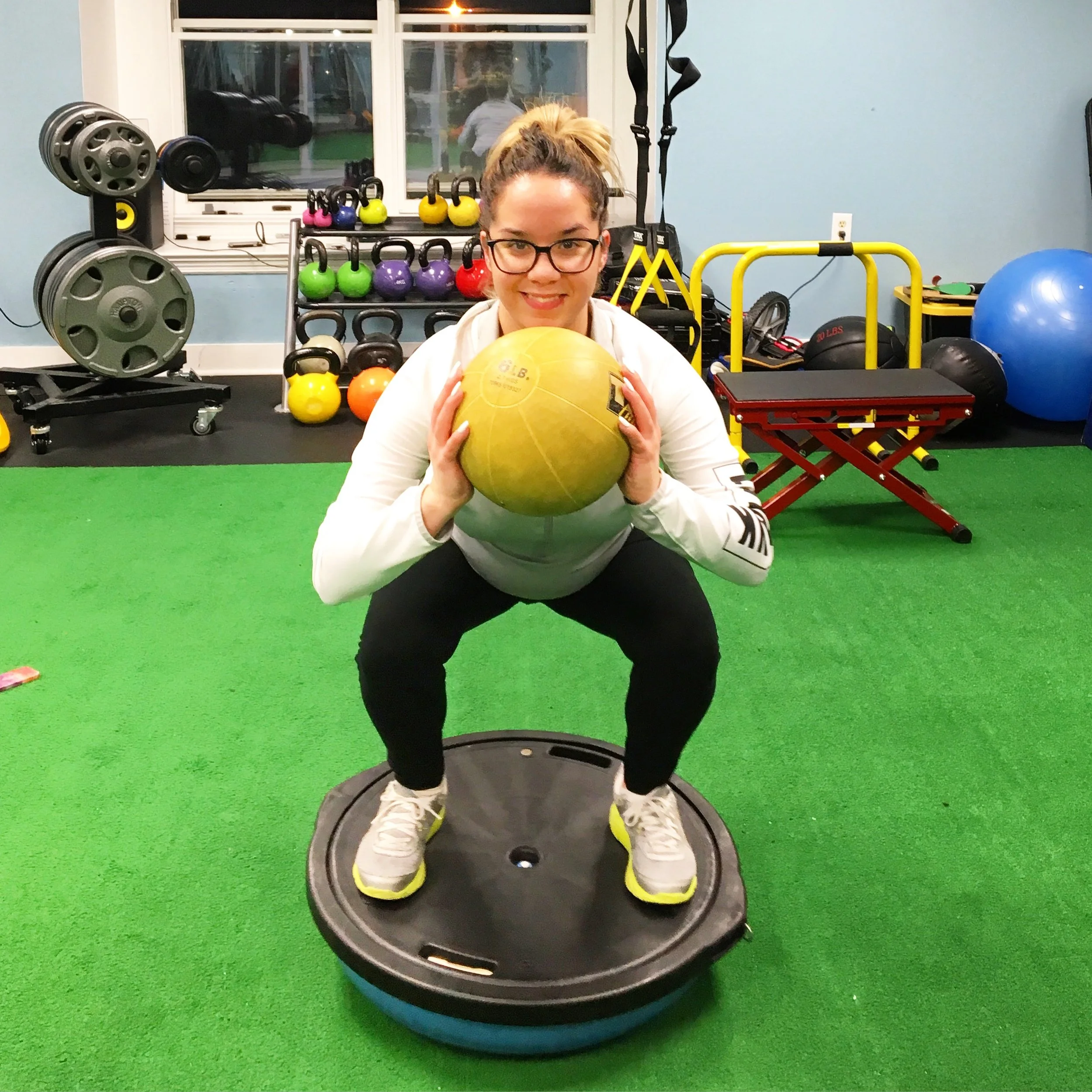 A woman with glasses and a high ponytail is kneeling on green turf, holding a yellow medicine ball. She is smiling. The room appears to be a fitness or therapy space with various exercise equipment, including kettlebells, weights, a blue exercise bal