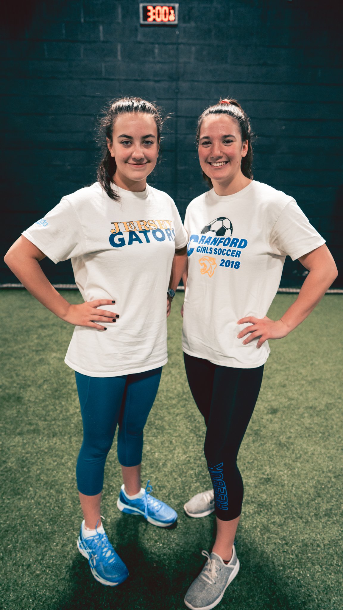 Two young women standing on an indoor soccer field, posing for a photo. They are wearing athletic clothes and smiling. The woman on the left is wearing a white T-shirt that says "Jersey Gators" and blue athletic leggings. The woman on the right is we
