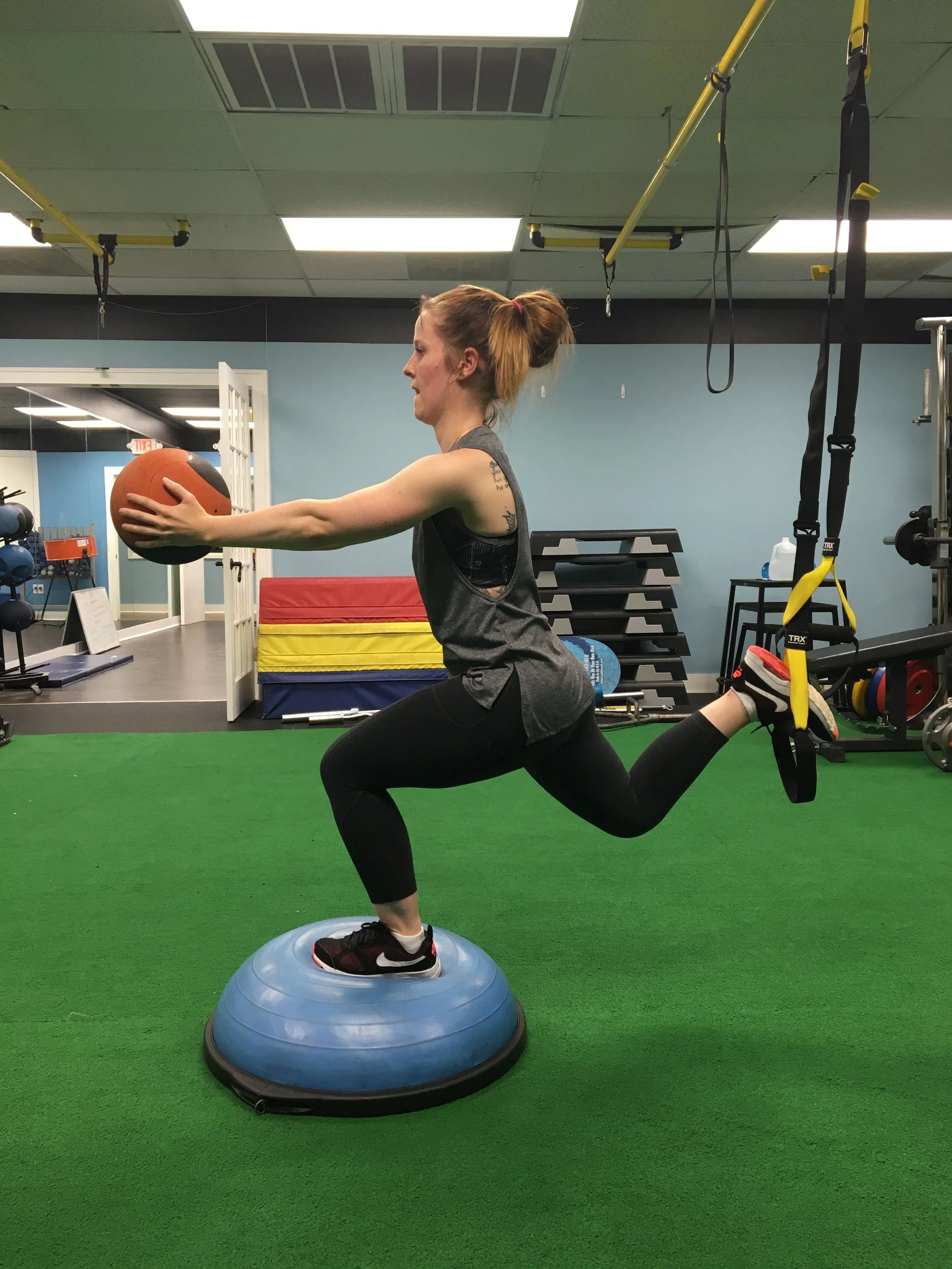 A woman performing a lunge exercise holding a medicine ball in a gym. She is balancing on a half-dome balance trainer with her right foot, with resistance bands hanging from the ceiling behind her.