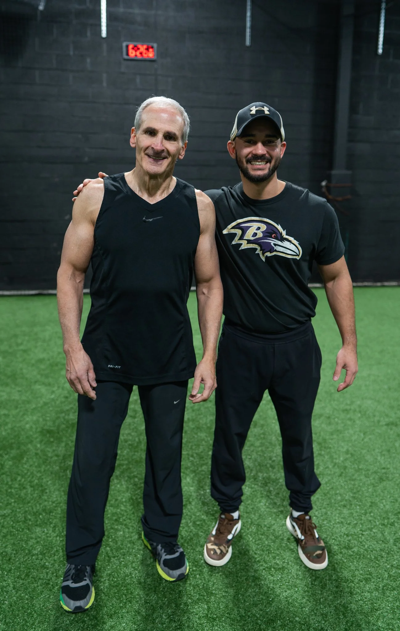 Two men standing on indoor turf, smiling, with the man on the left wearing a sleeveless athletic shirt and the man on the right wearing a T-shirt with a Baltimore Ravens logo and a baseball cap, posing with an arm around each other's shoulders.