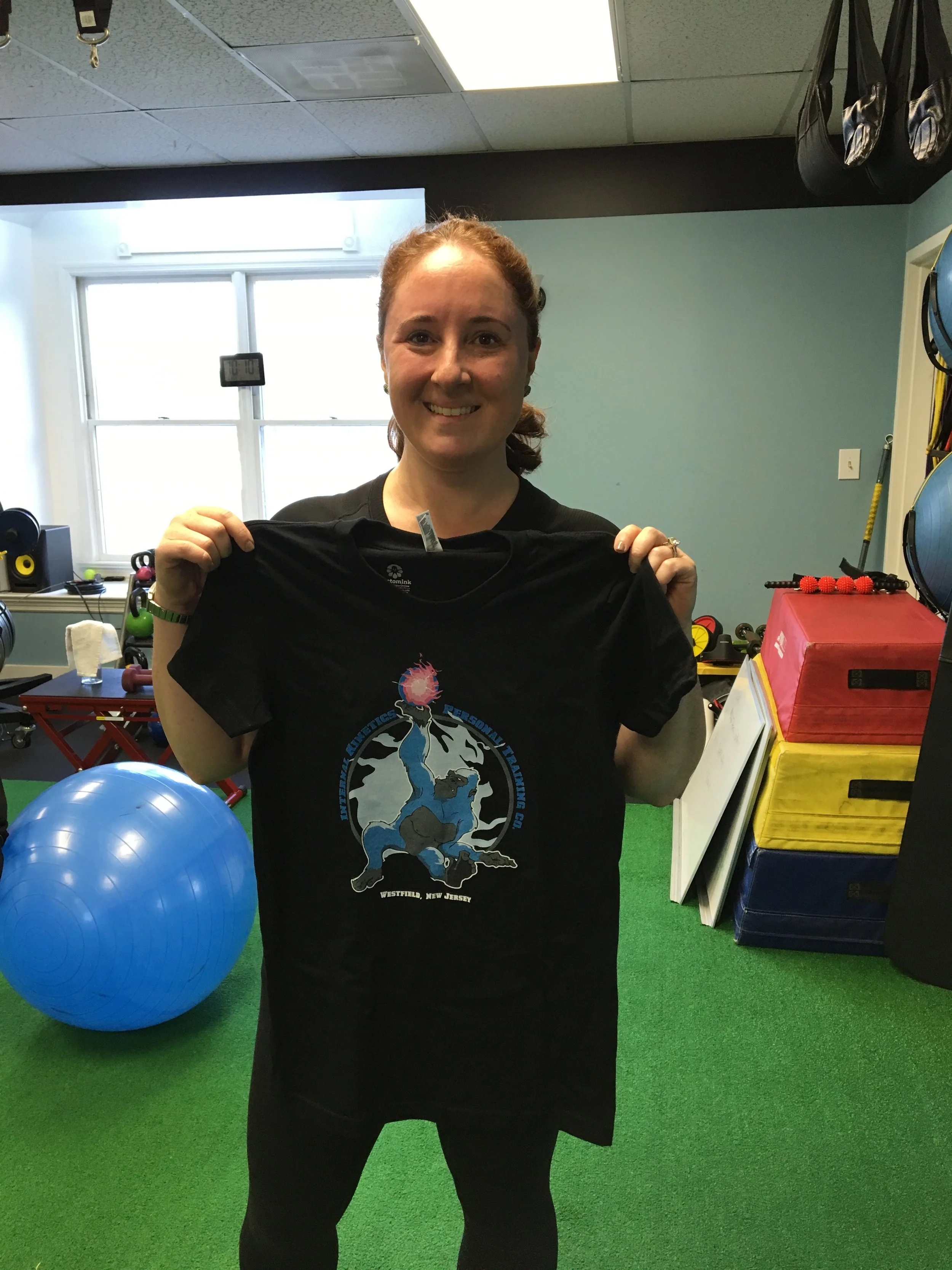A woman with red hair holding a black T-shirt in a gym. There is a large blue exercise ball in front of her, and gym equipment including weights and boxes in the background.
