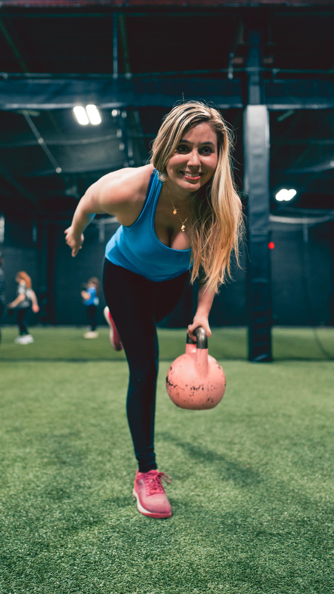 A woman in a blue tank top and pink sneakers exercising with a kettlebell in a gym inside a fitness center.
